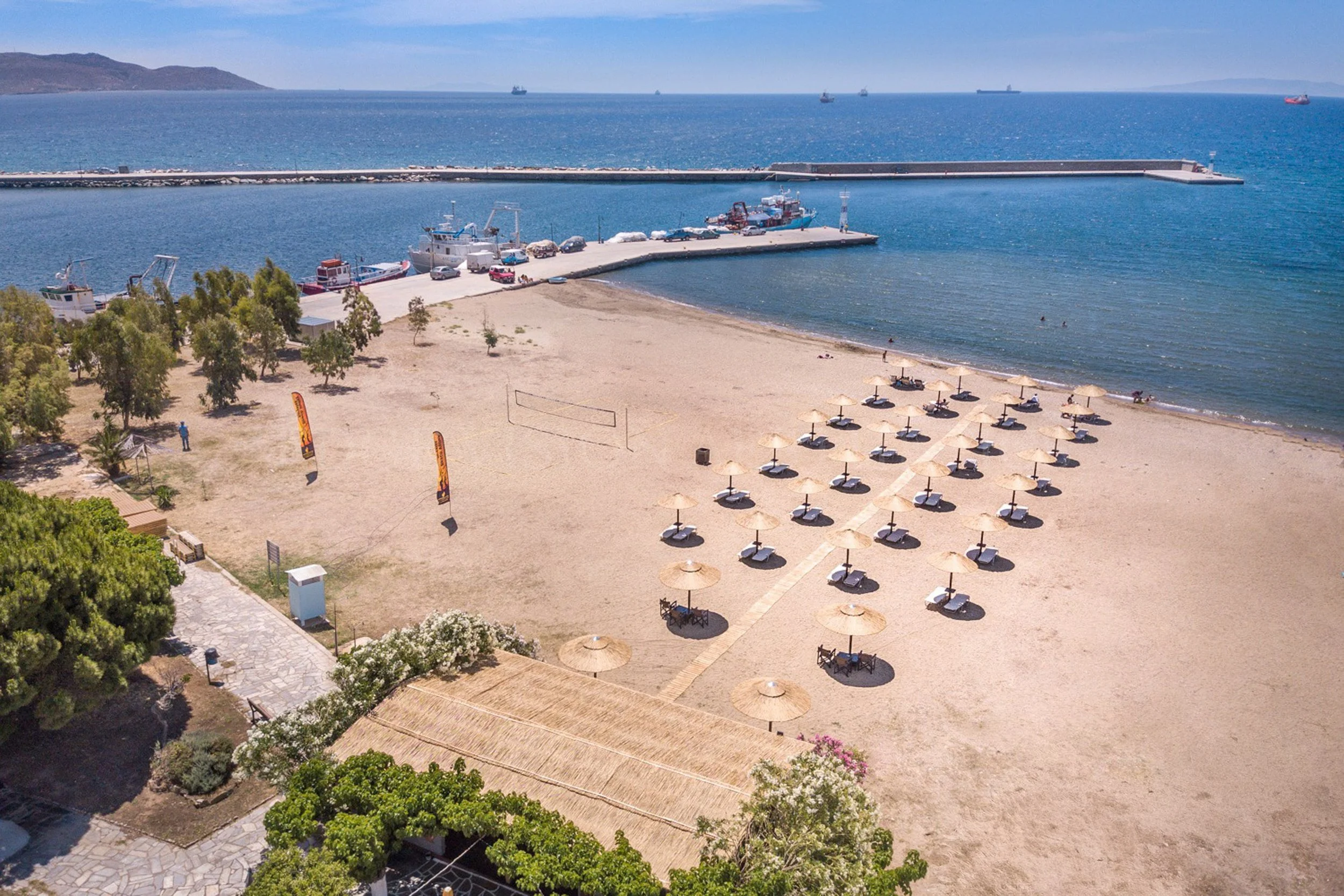 A beach with rows of umbrellas and lounge chairs near the shoreline, a sandy area, and a small pier with boats, beyond which blue water and ships are visible, with distant mountains on the horizon.