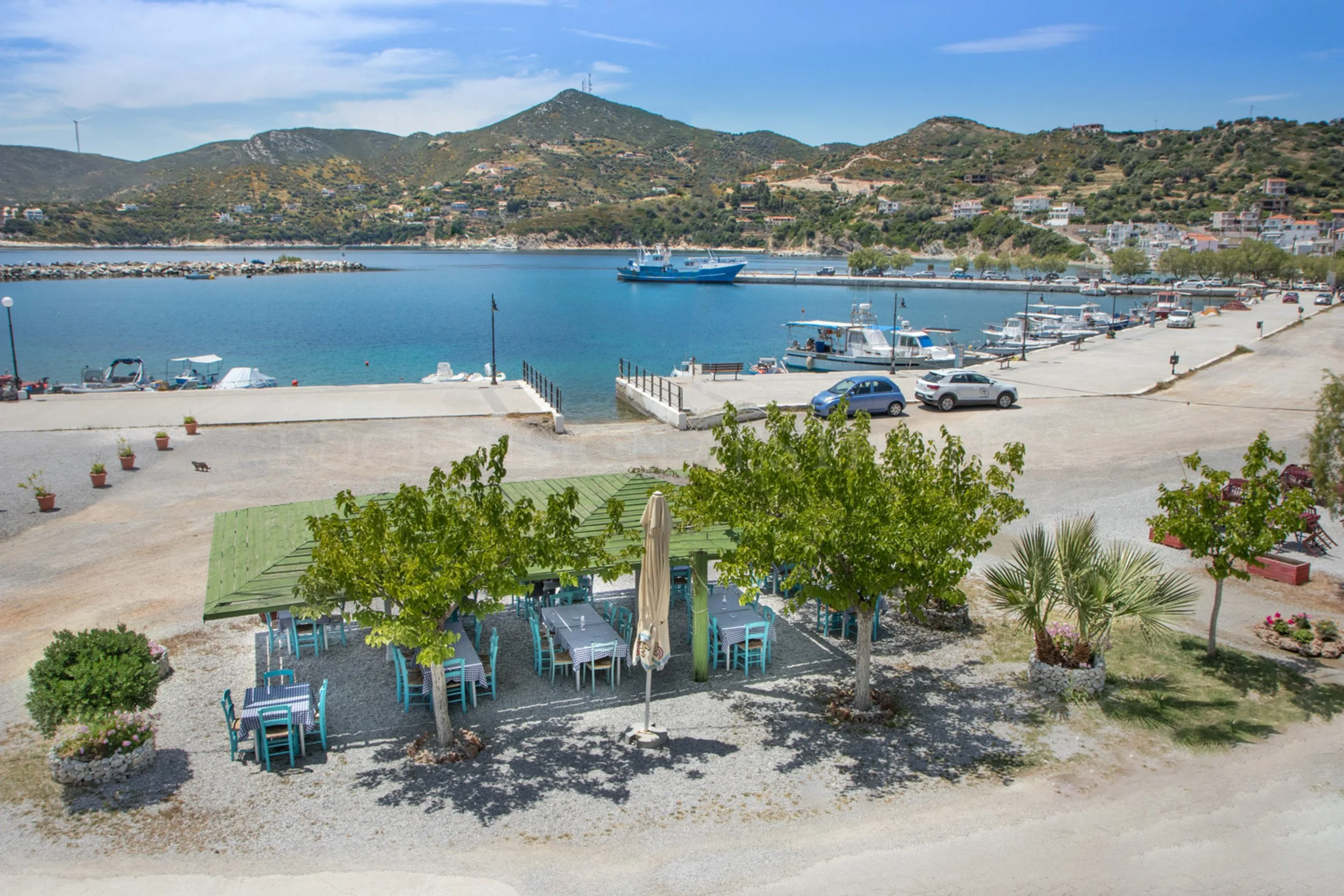 Outdoor cafe with blue chairs and tables under green umbrellas, near a marina with boats, surrounded by trees and mountains in the background.