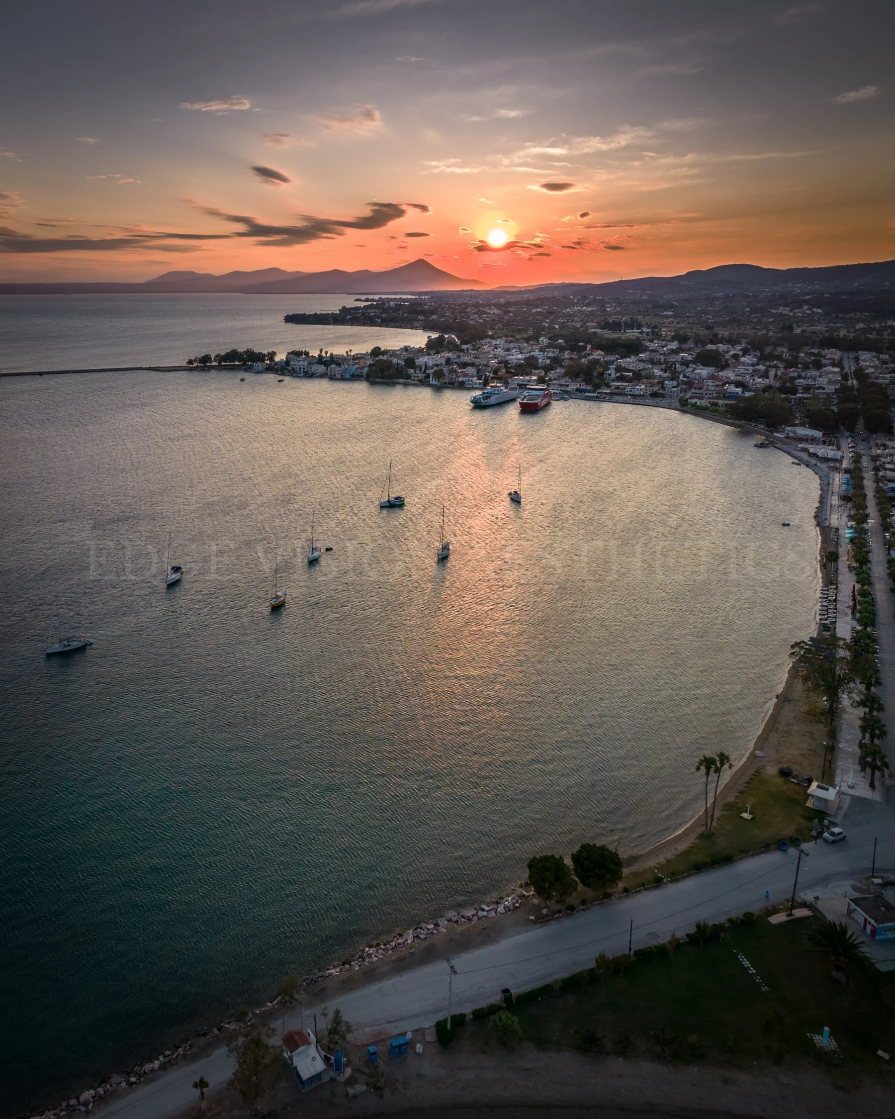 Aerial view of a coastal town at sunset with sailboats in the water, a shoreline lined with trees, and mountains in the background.