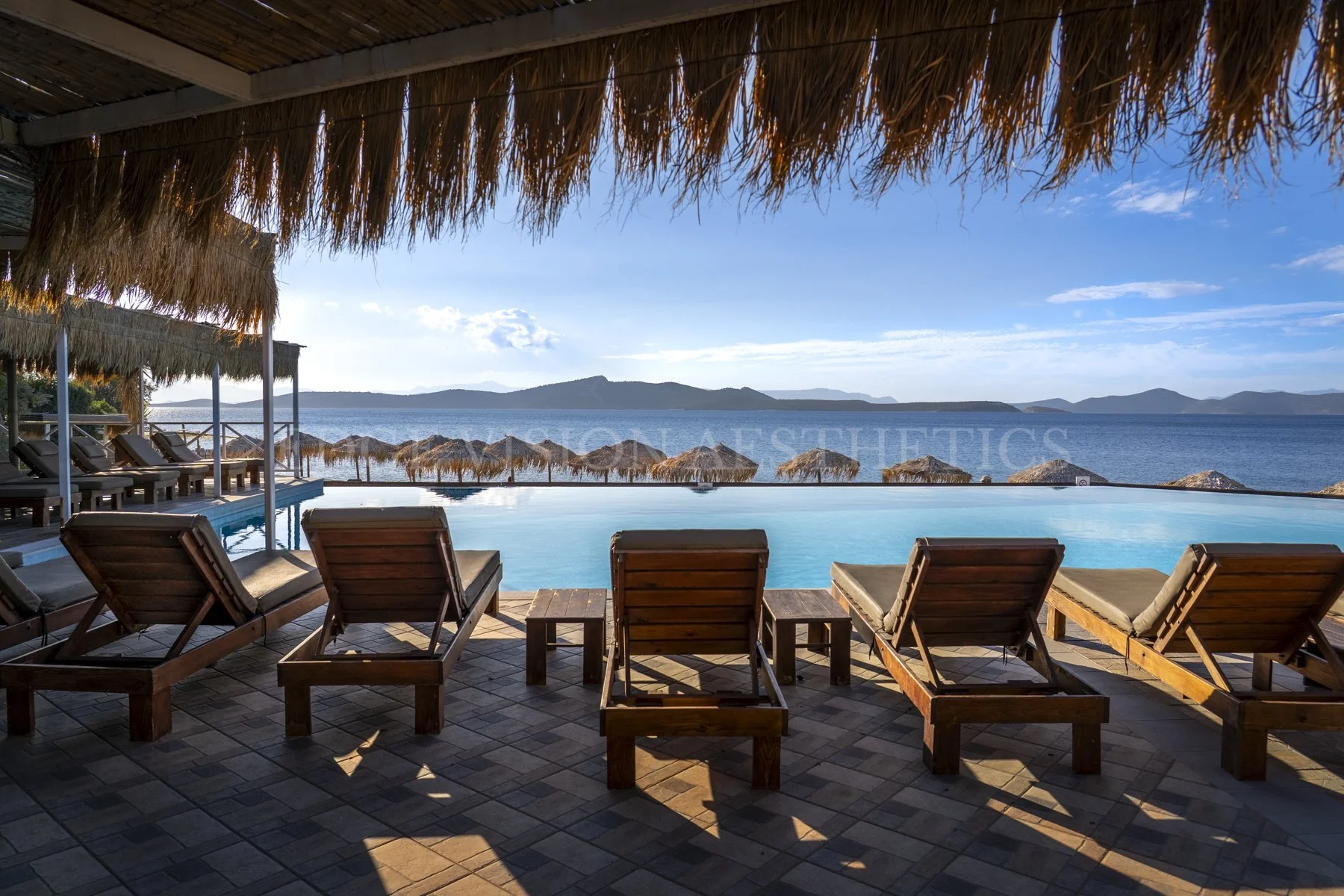 Wooden lounge chairs and small tables on a shaded deck overlooking an infinity pool with a view of the ocean, islands in the distance, and a clear blue sky with a few clouds.