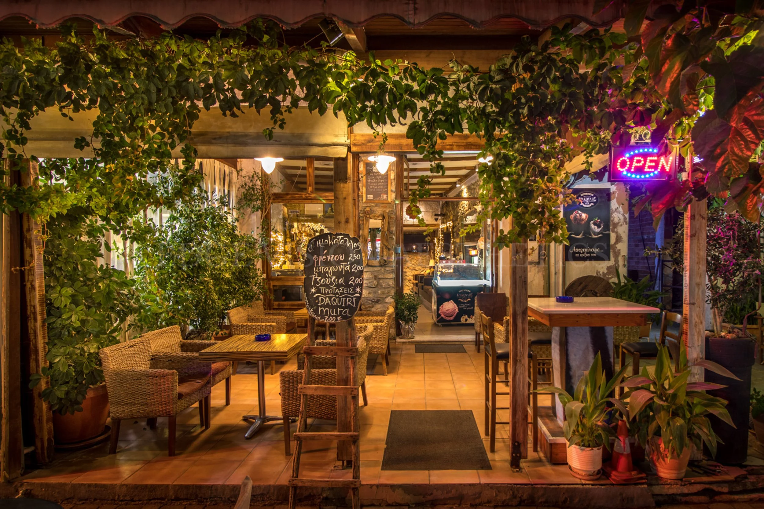 Night view of a cozy outdoor restaurant with wooden furniture, string lights, and lush potted plants, including a neon 'OPEN' sign.