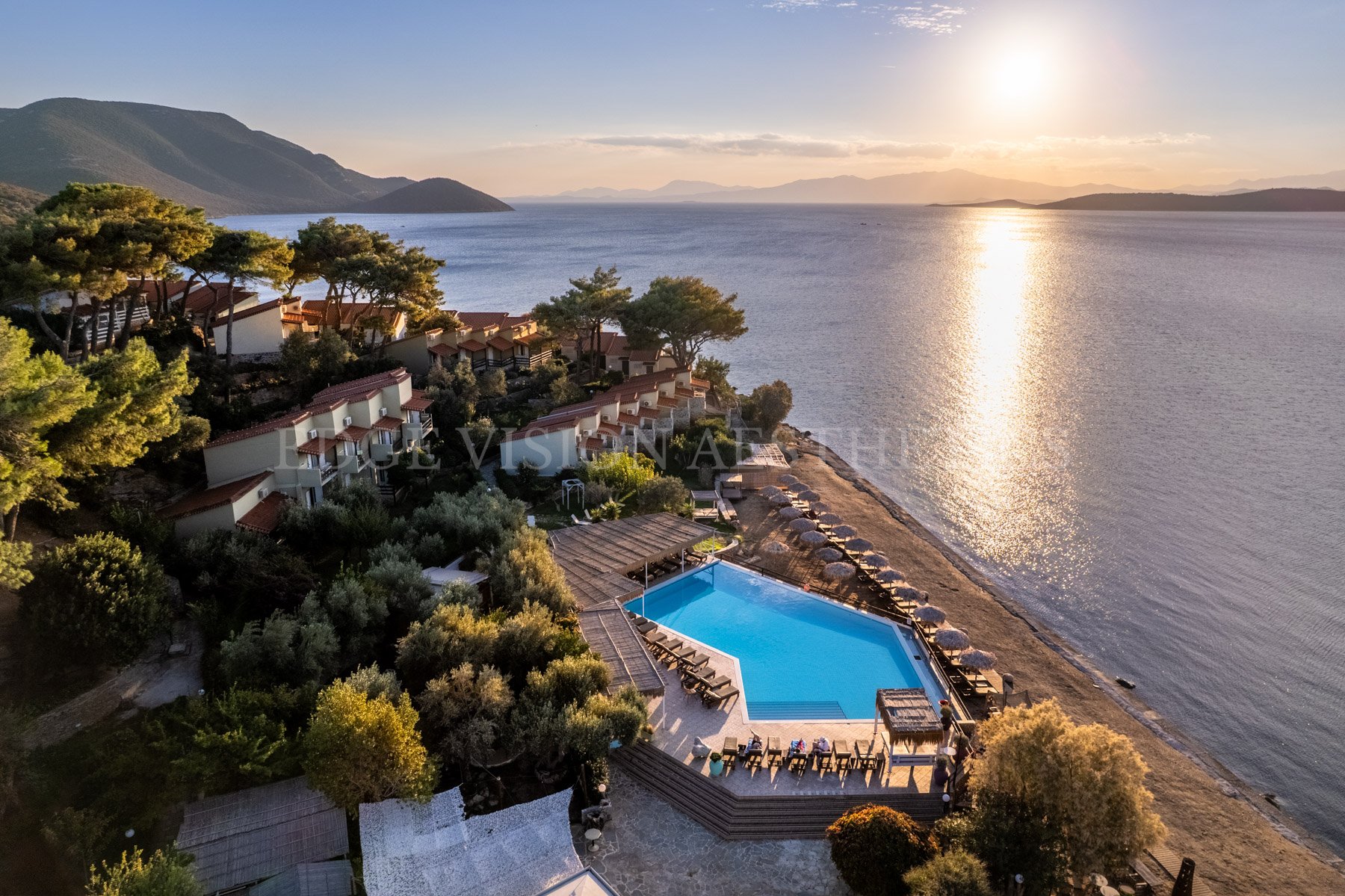 Aerial view of a seaside resort at sunset, featuring a swimming pool, beach umbrellas, trees, hillside houses, and calm water reflecting the setting sun with mountains in the background.