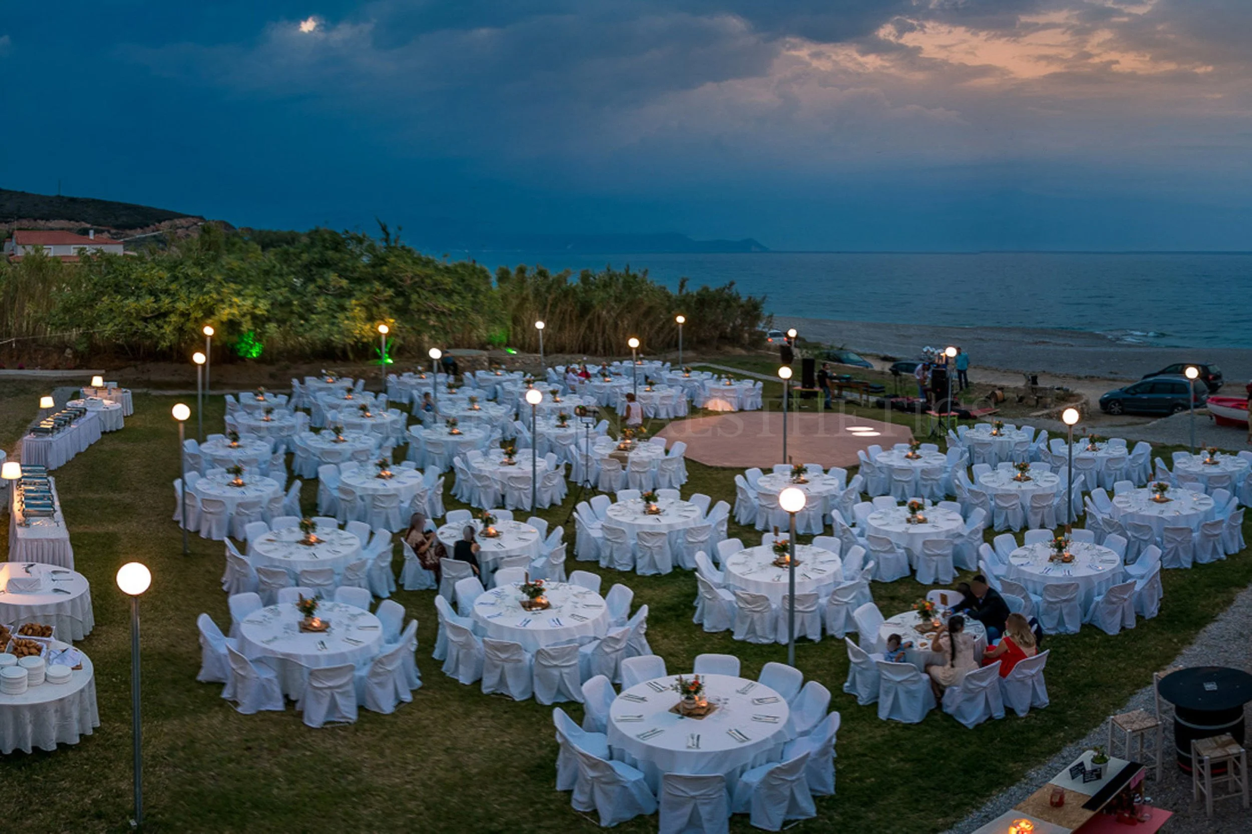 Outdoor dining setup with round tables covered in white tablecloths, set with plates and candles, along a beach with water and a cloudy sky in the background, during dusk.