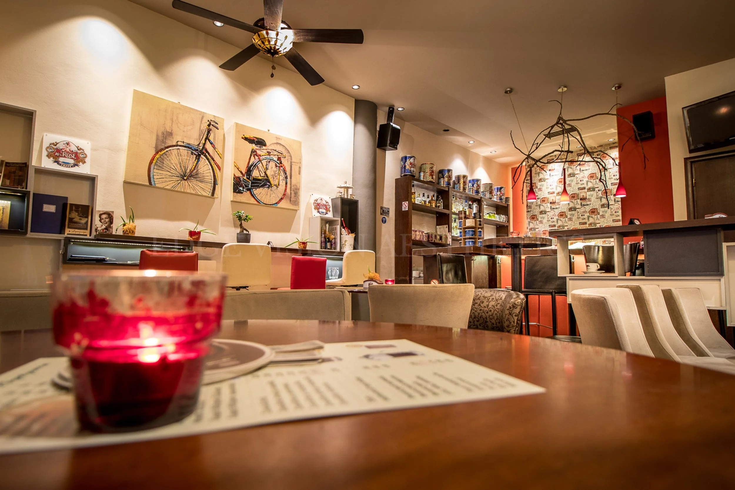 Inside a cozy cafe or restaurant with tables, chairs, artwork of bicycles on the wall, and decorative branch lighting fixtures. The foreground shows a red candle holder and a menu, with a blurred view of the interior in the background.