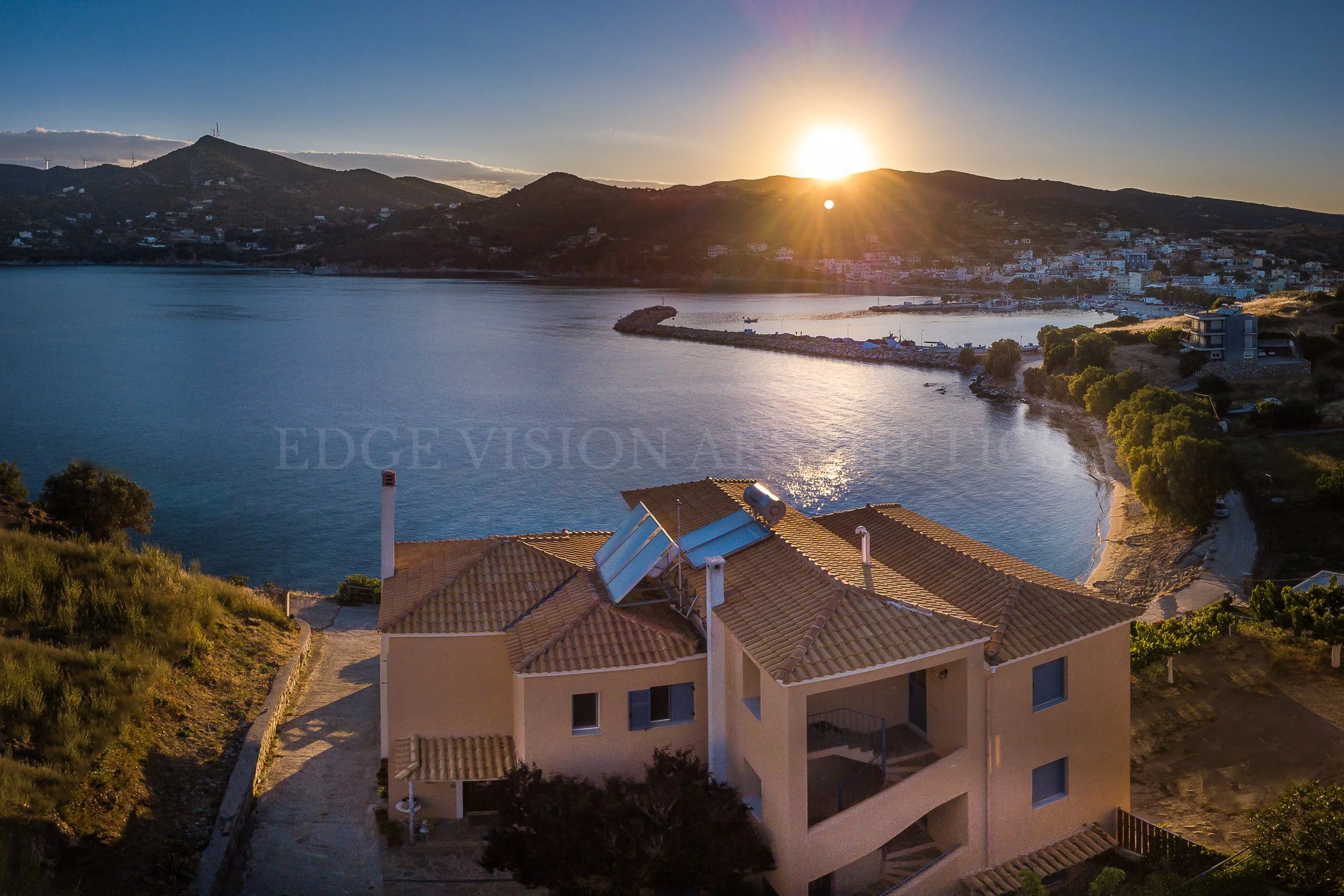 A scenic view of a coastal town at sunset, with houses on a hillside, a body of water, and the sun setting behind the mountains.