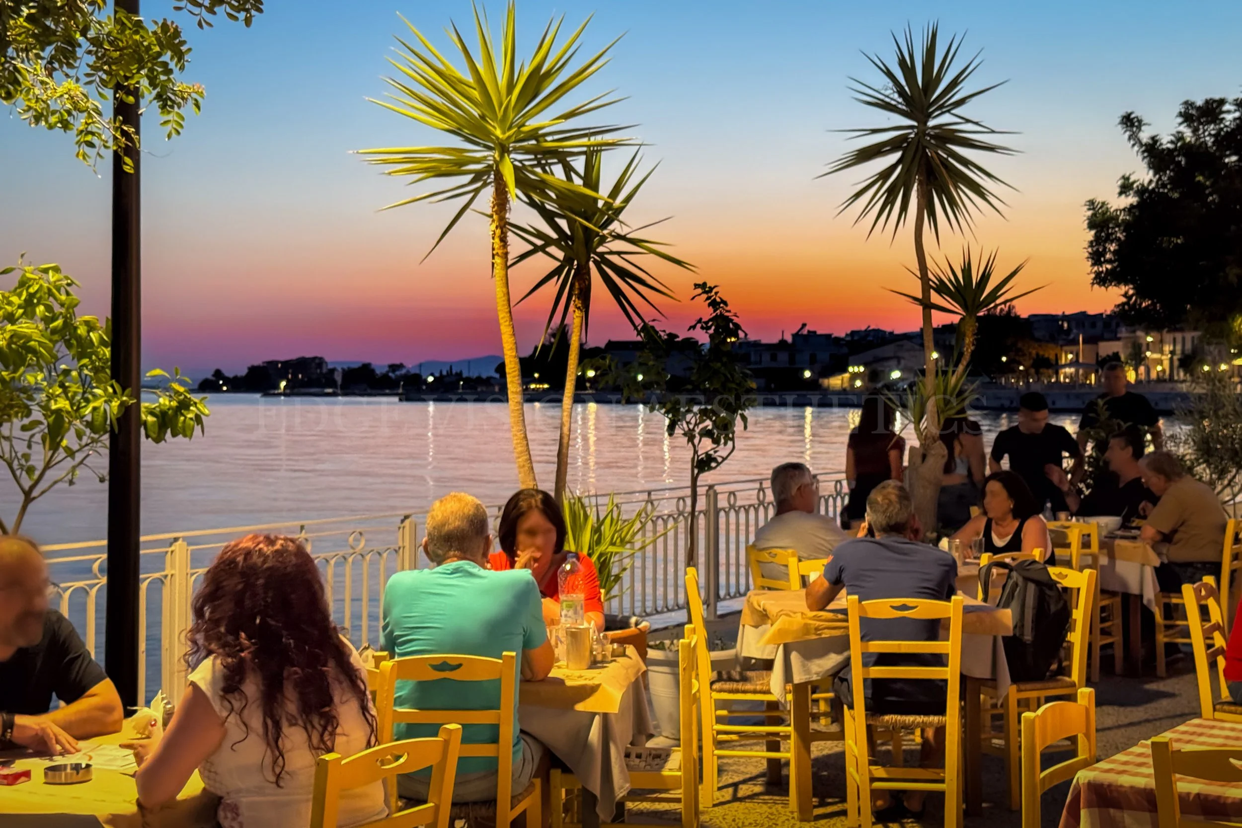 People dining outdoors at a restaurant by the water during sunset, with tall spiky palm-like trees in the background and colorful sky.