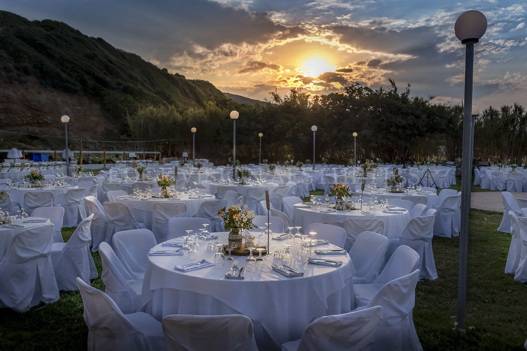 Outdoor event setup with round tables covered in white tablecloths, decorated with floral centerpieces, set with glasses and utensils, arranged on a grassy area during sunset, with a backdrop of hills and cloudy sky.
