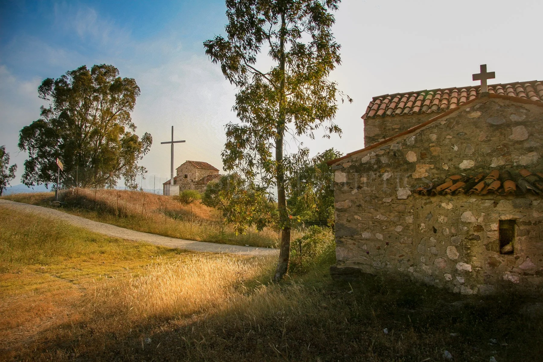 A rural scene with an old stone church with a tiled roof and a cross on top, surrounded by trees and a grassy area with a dirt path, under a mostly clear sky