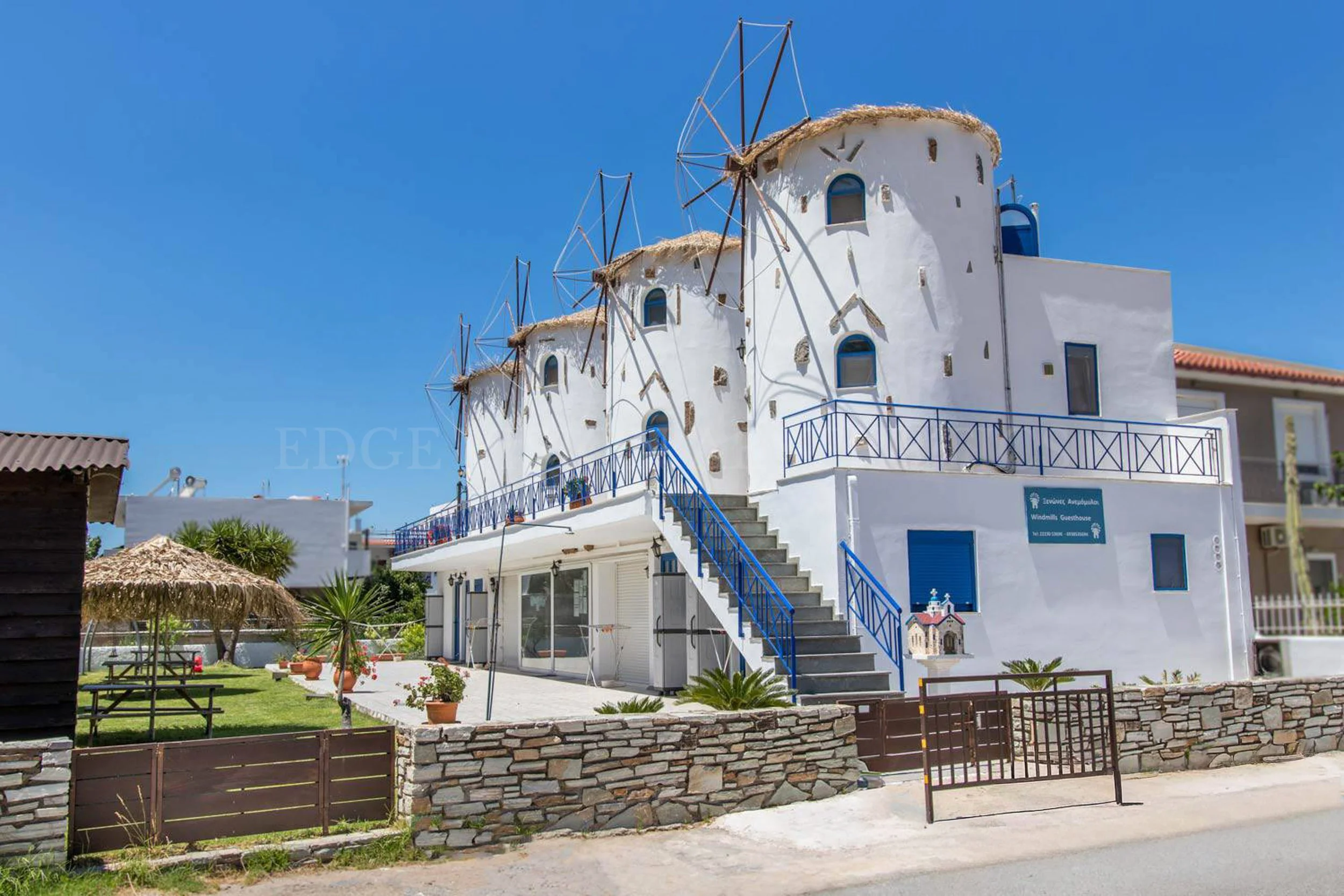 A white building with blue accents, multiple windmill structures on the roof, blue staircases, and a sign that reads "Eretria Autobuses Windmills Guesthouse". There are potted plants, a small patio, a garden area with picnic tables, and a stone fence