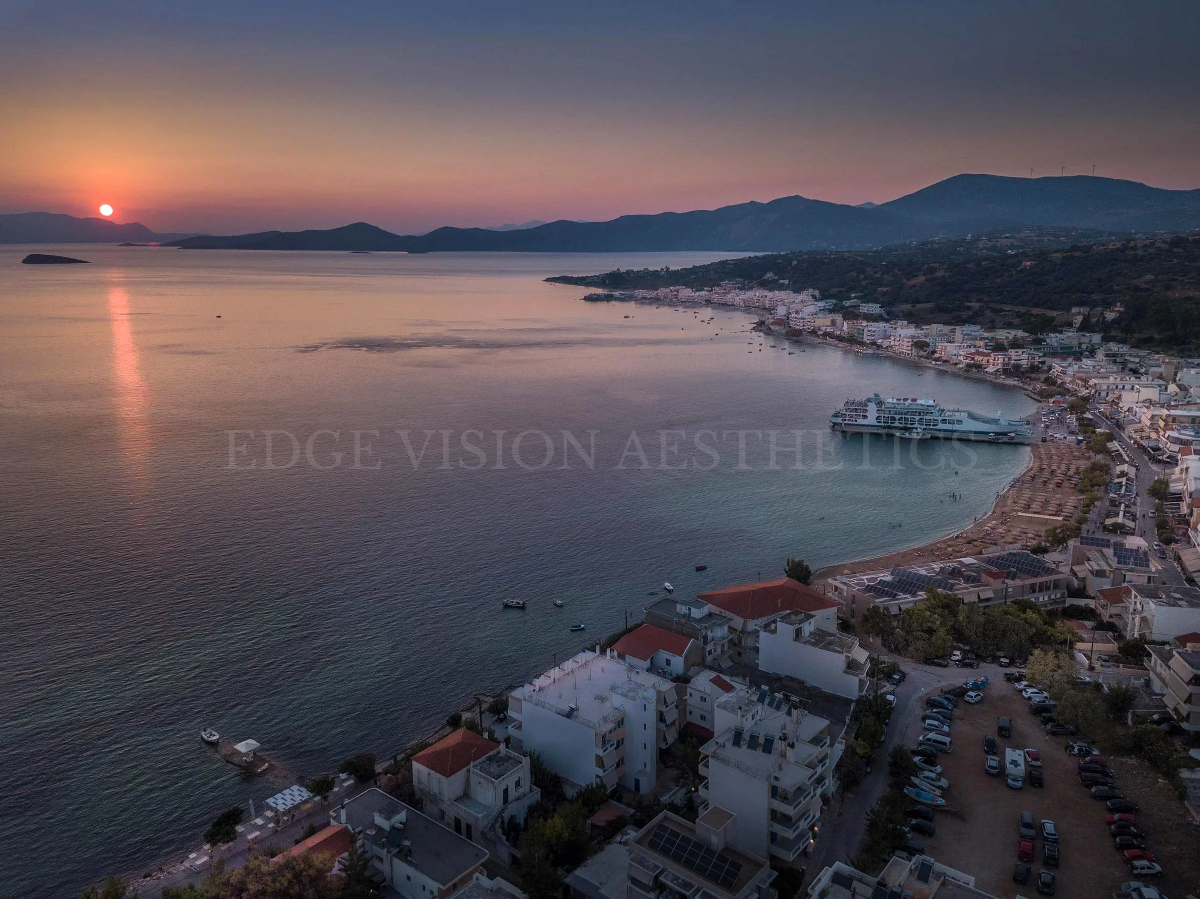 Aerial view of a coastal town at sunset with calm water, mountains in the background, and buildings along the shoreline.