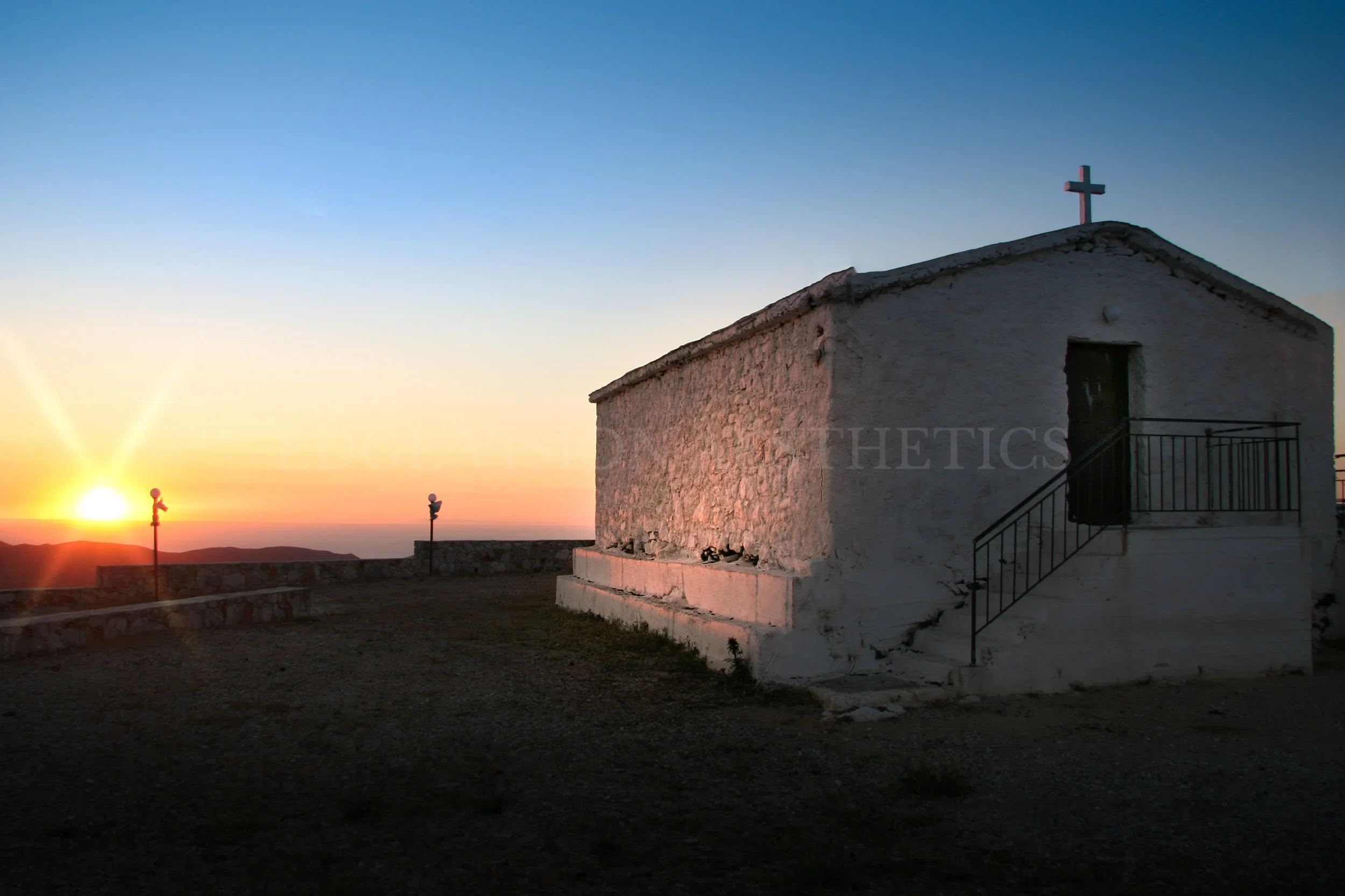 A small white chapel with a cross on top, set against a sunset sky with a bright sun low on the horizon, distant hills, and a few street lamps in the area.