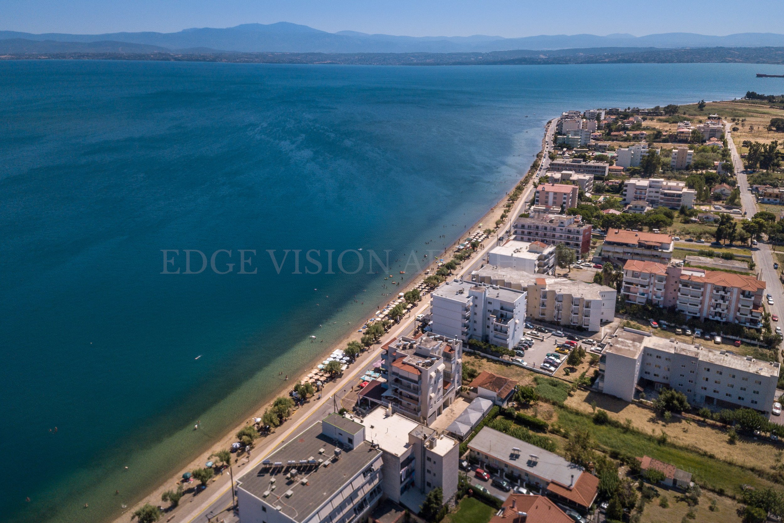 Aerial view of a coastal city with a beach, high-rise buildings, and a large body of water, with mountains in the background.