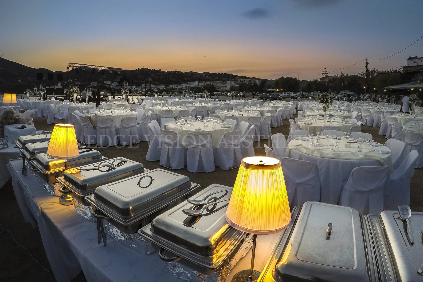 Outdoor wedding reception setup at sunset with round tables covered in white cloth, white chairs, floral centerpieces, buffet chafing dishes, and yellow table lamps.
