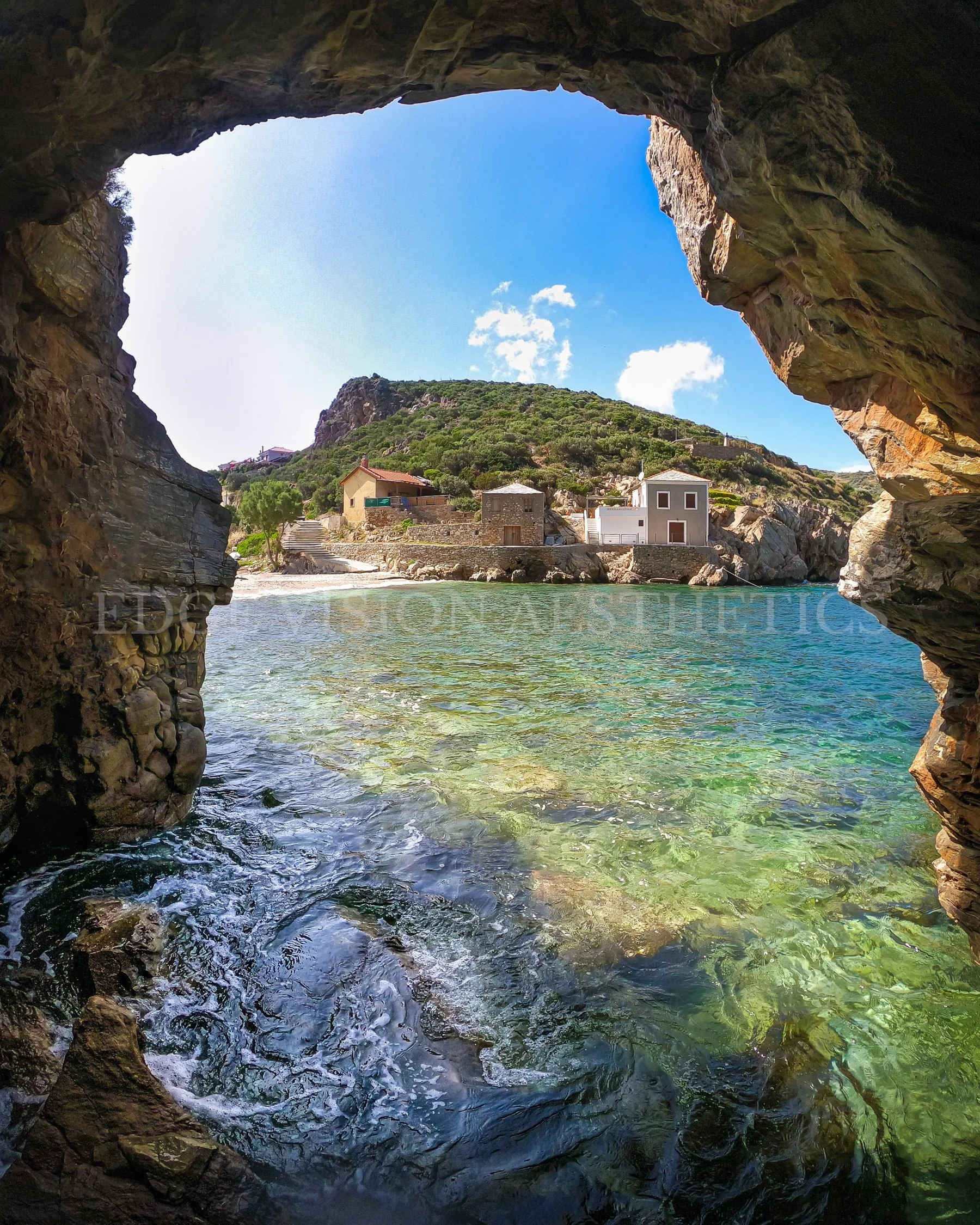 View from inside a rocky cave looking out towards bright blue sky, green hillside, houses, and water, with sunlight illuminating the scene.