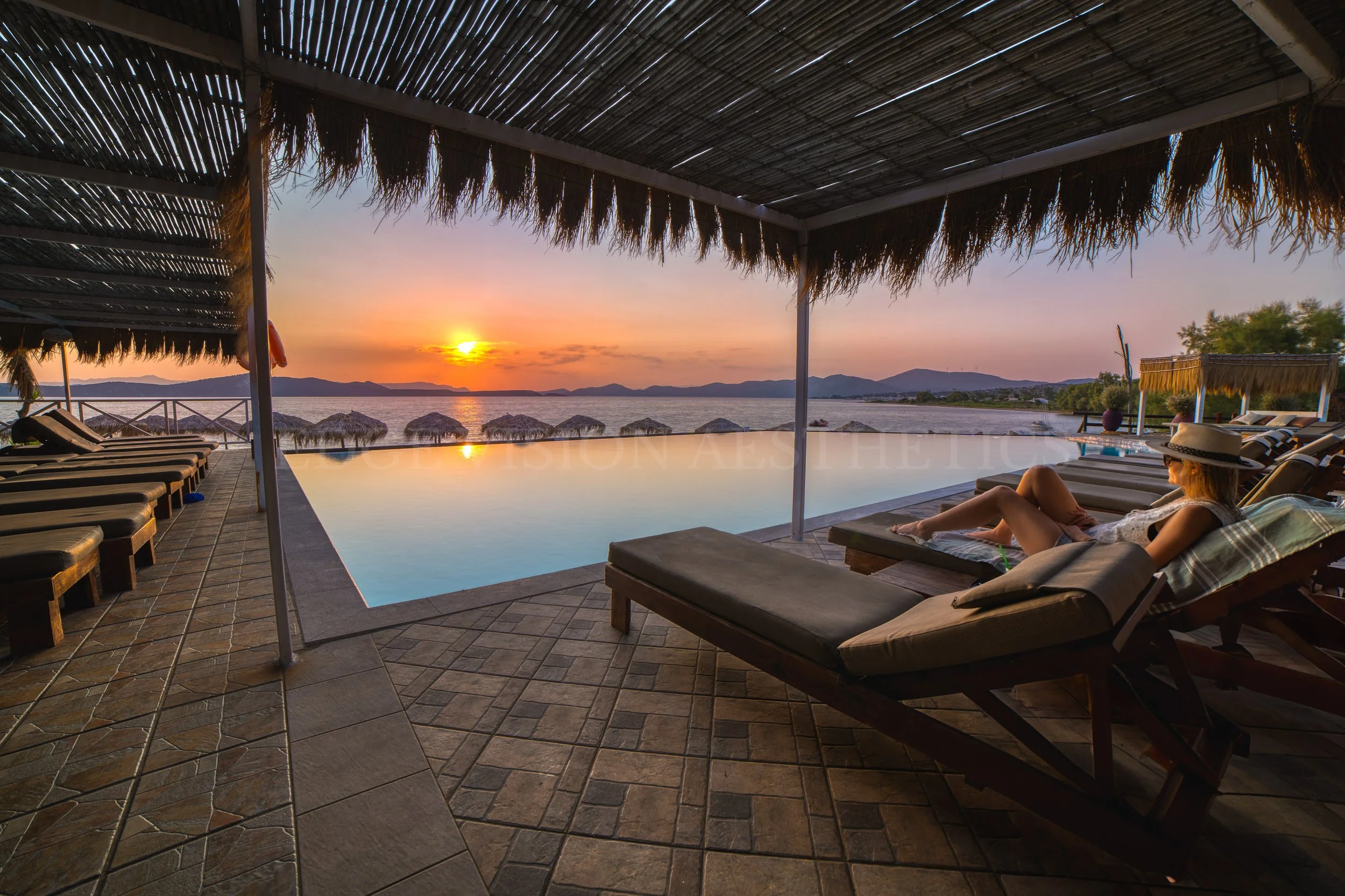 Resort pool area with lounge chairs under a thatched roof cabana overlooking a sunset over the water and mountains.