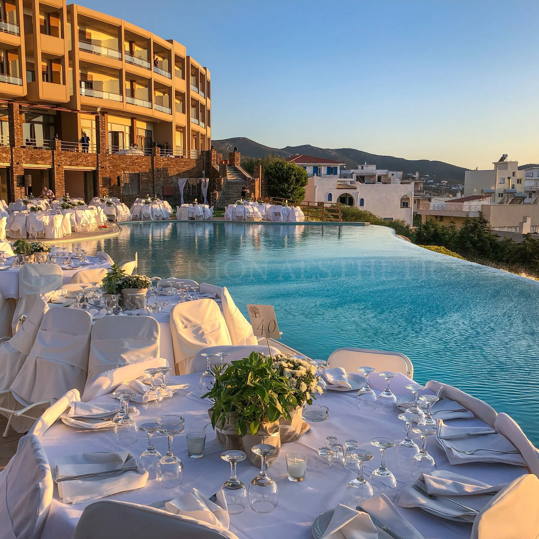 Elegant outdoor dining setup by a swimming pool with white tablecloths, floral centerpieces, and glassware, overlooking a scenic hillside with residential buildings during sunset.