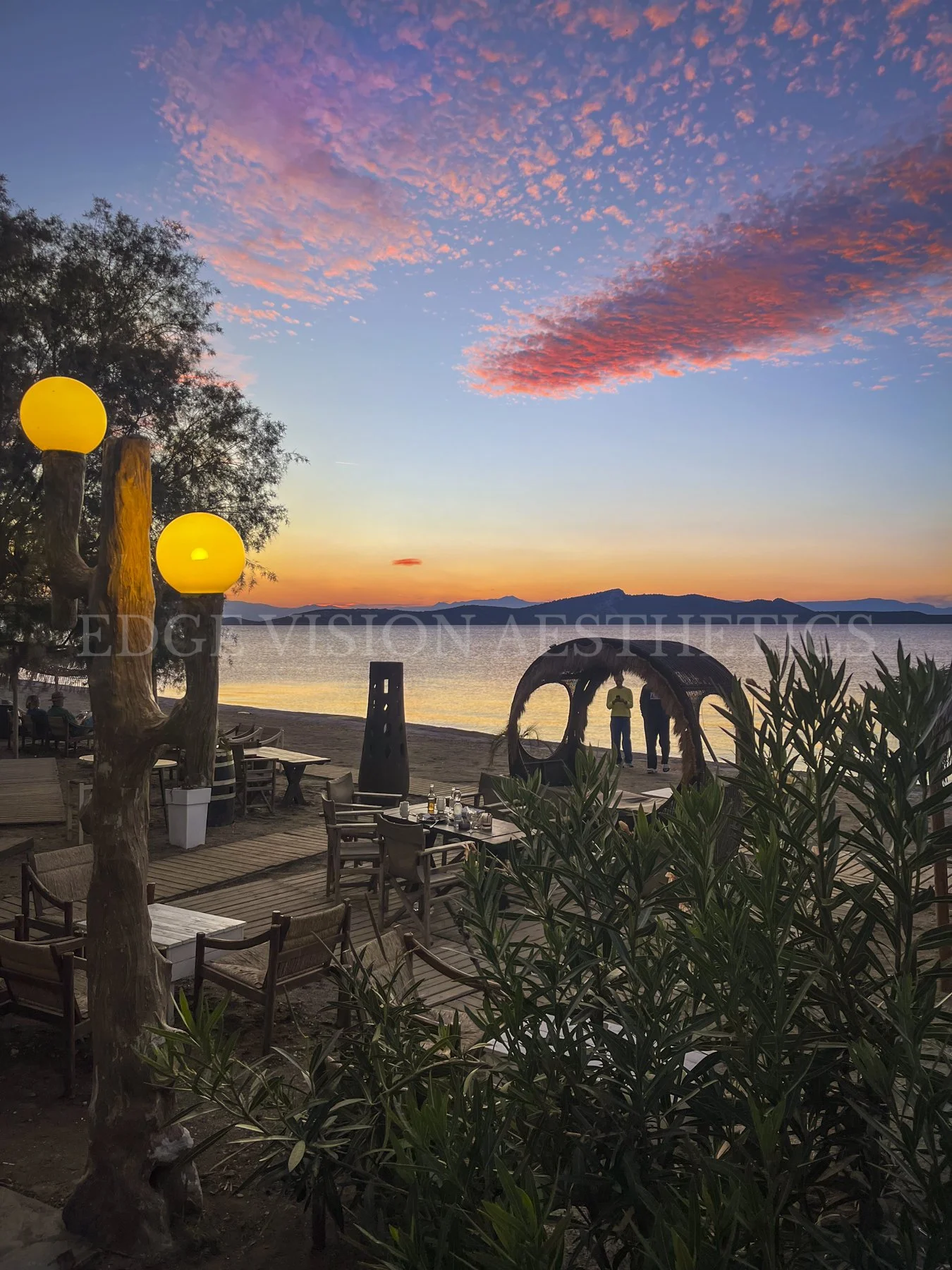A lakeside restaurant or bar at sunset with outdoor seating, wooden decking, trees, and pink and orange clouds in the sky.