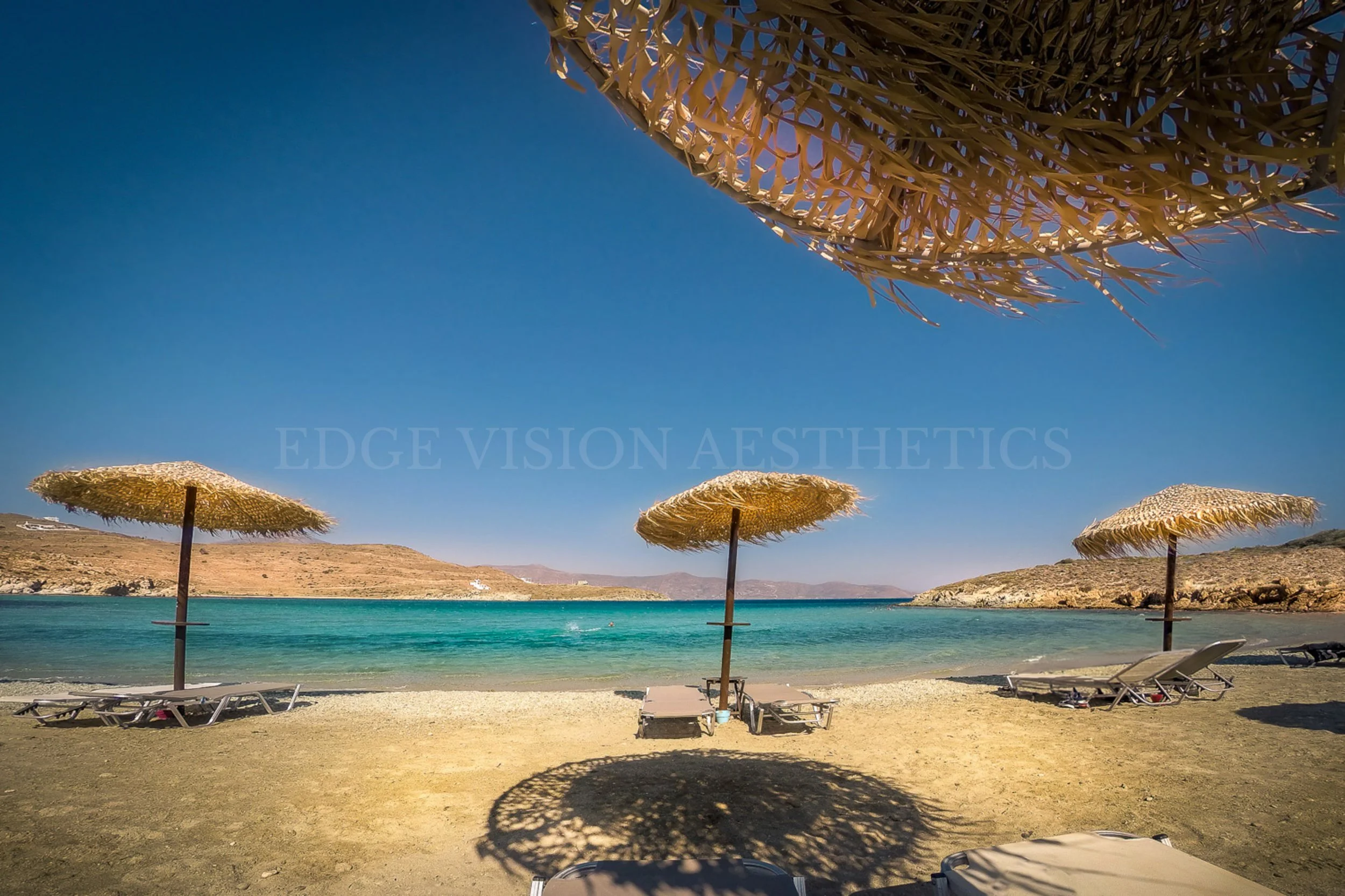 Beach scene with straw umbrellas and lounge chairs by the water under a clear blue sky.