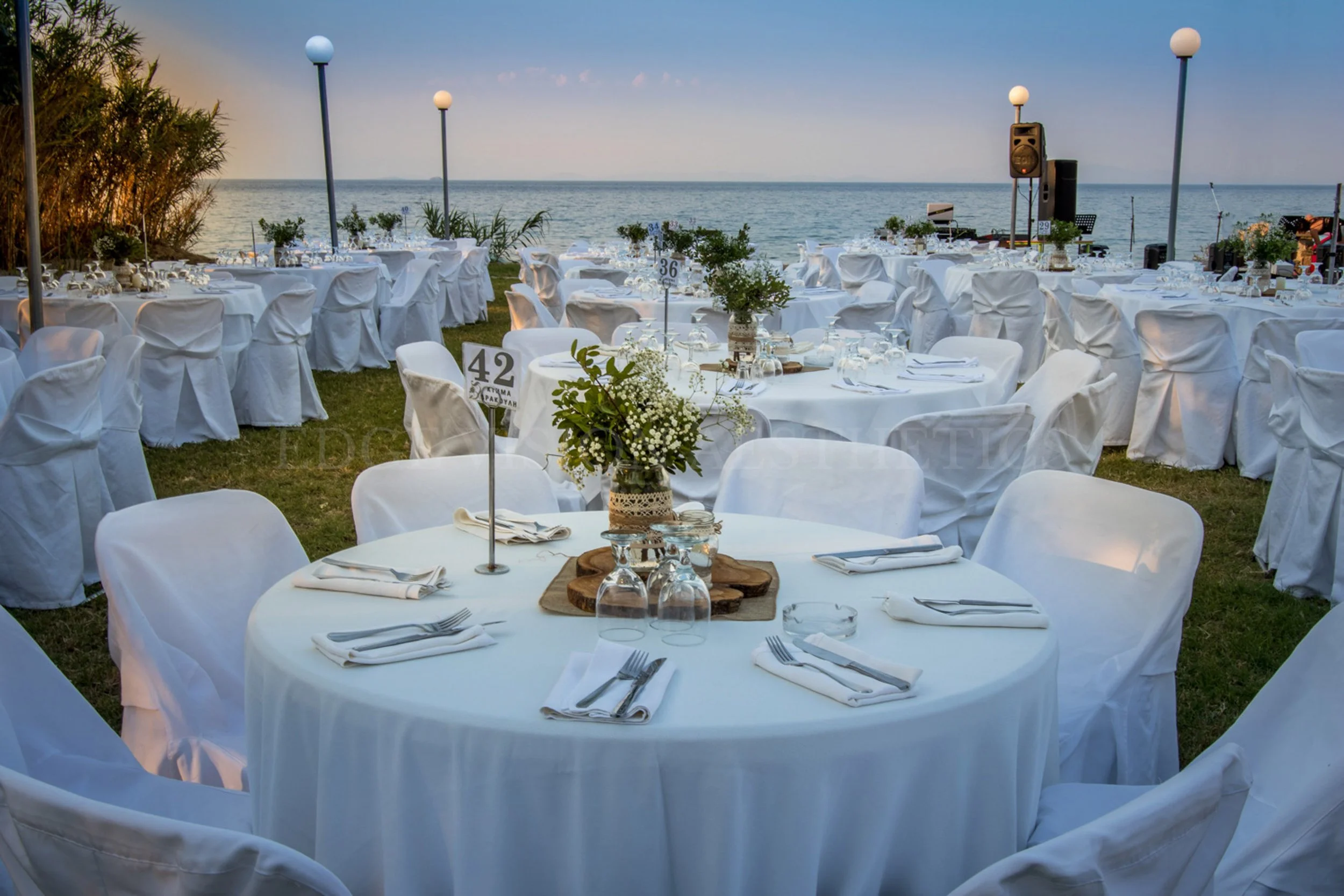Wedding reception setup outdoors by the water with round tables covered in white tablecloths, decorated with floral centerpieces and table numbers, and surrounded by white chairs, with lamps and a speaker system, during sunset