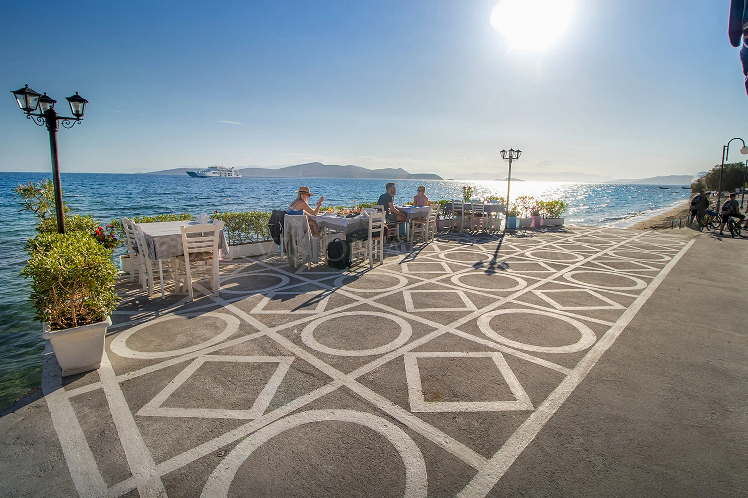Outdoor dining area by the sea with white chairs and tables, decorative black lattice pavement, potted plants, vintage-style street lamps, and a distant ferry boat on the water, under clear blue sky and bright sunlight.