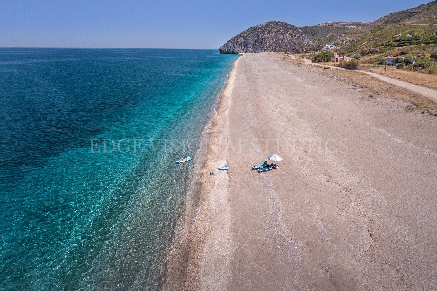 Drone view of a quiet, sandy beach with a few people relaxing on lounge chairs under a large umbrella, near the clear blue water, with a mountainous landscape in the background.