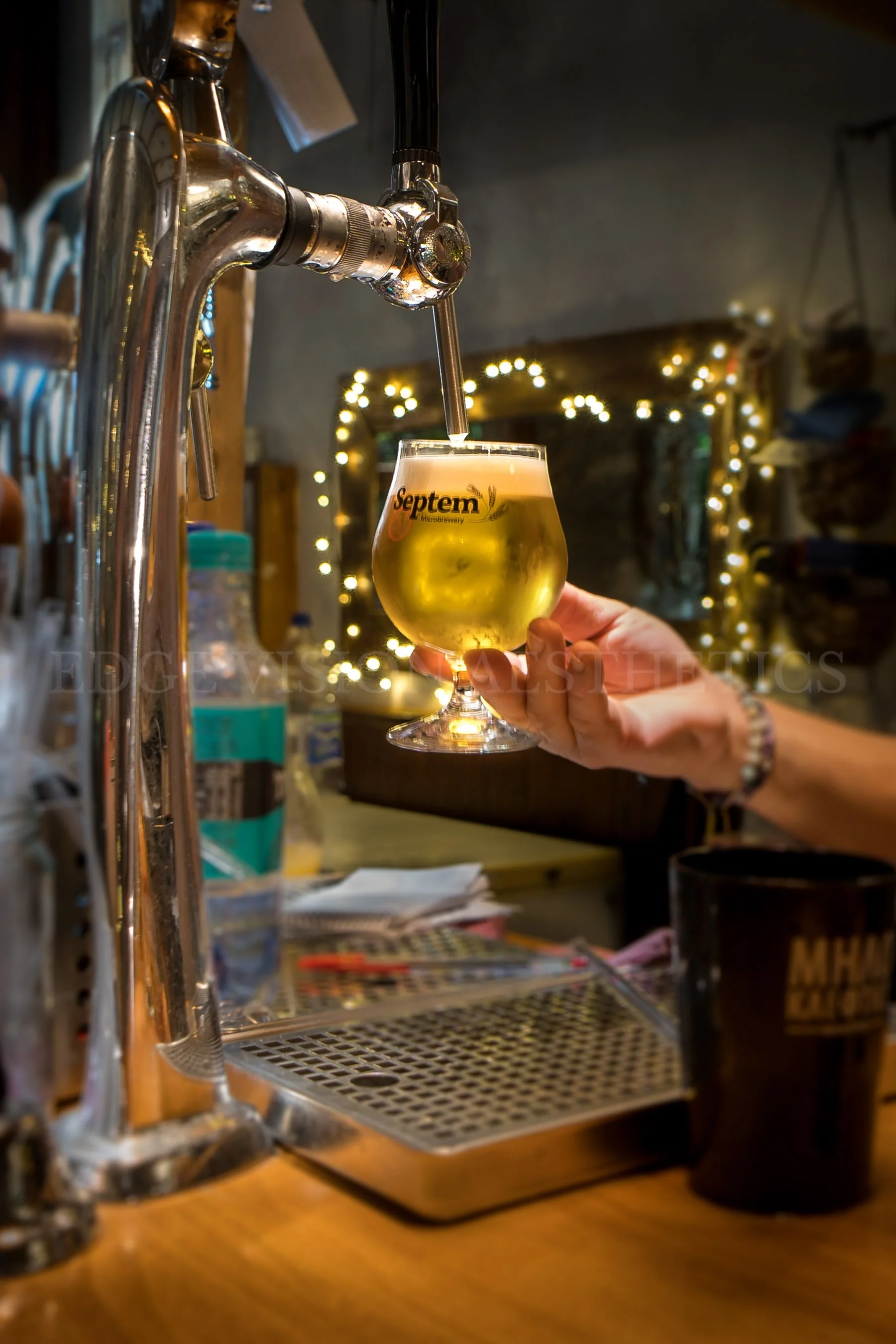 A hand holding a glass of beer under a beer tap, with a decorated and illuminated background.