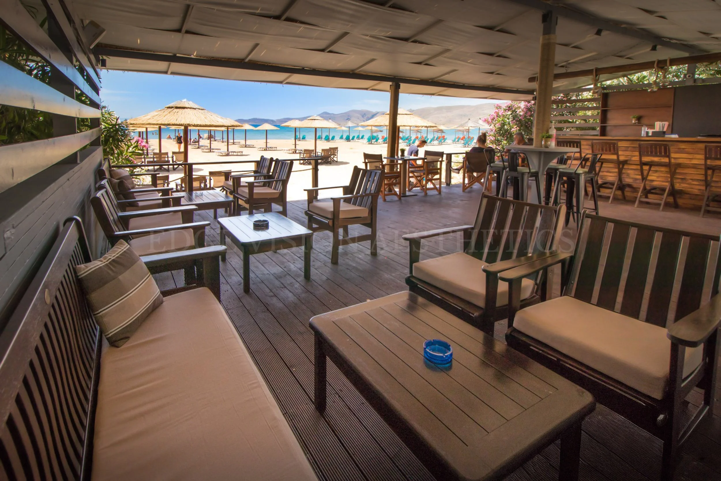 Beachside restaurant with outdoor seating, umbrellas, and lounge chairs by the ocean, mountains in the background.
