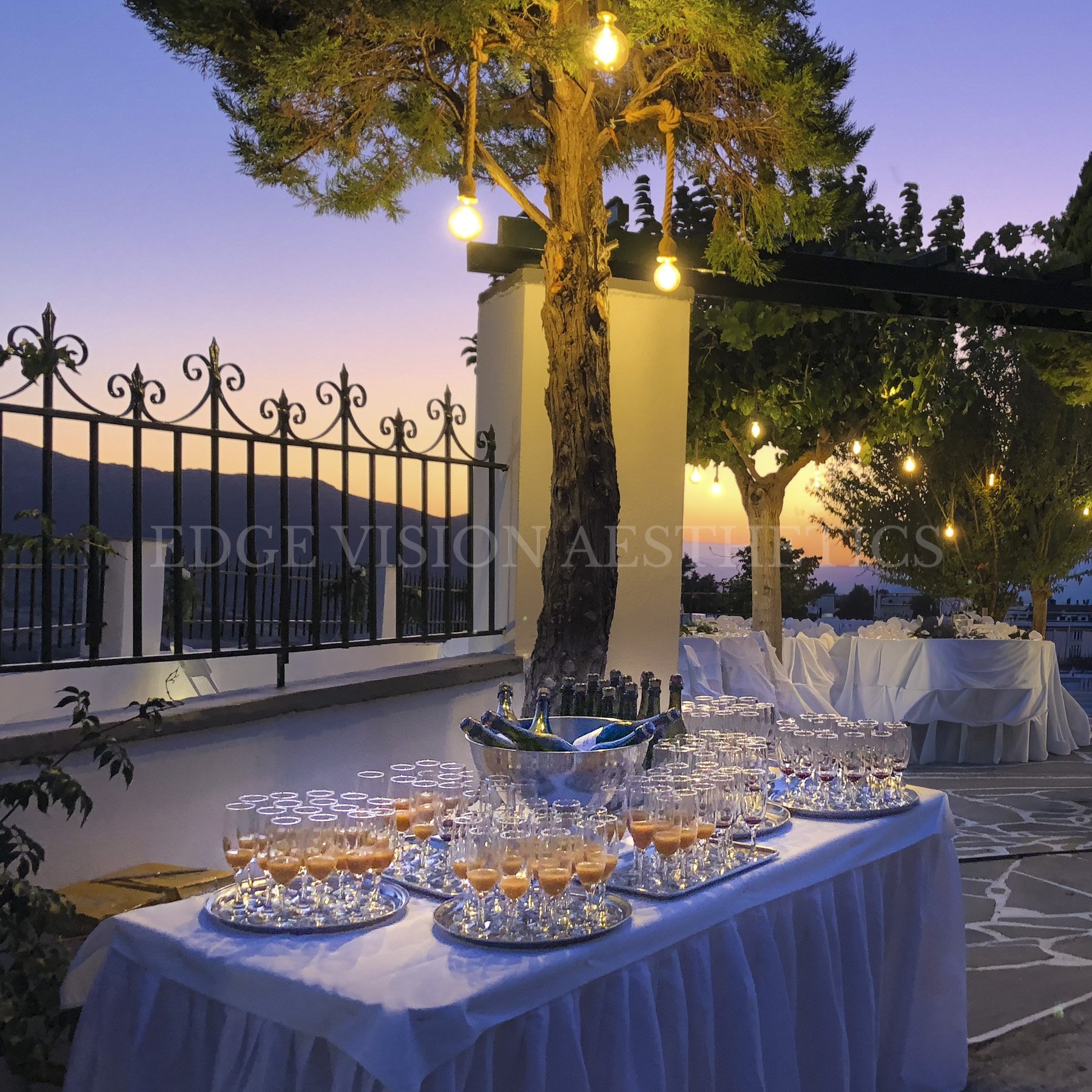 An outdoor sunset setting with trees decorated with hanging string lights, a white fencing, and a table with glasses and appetizers.
