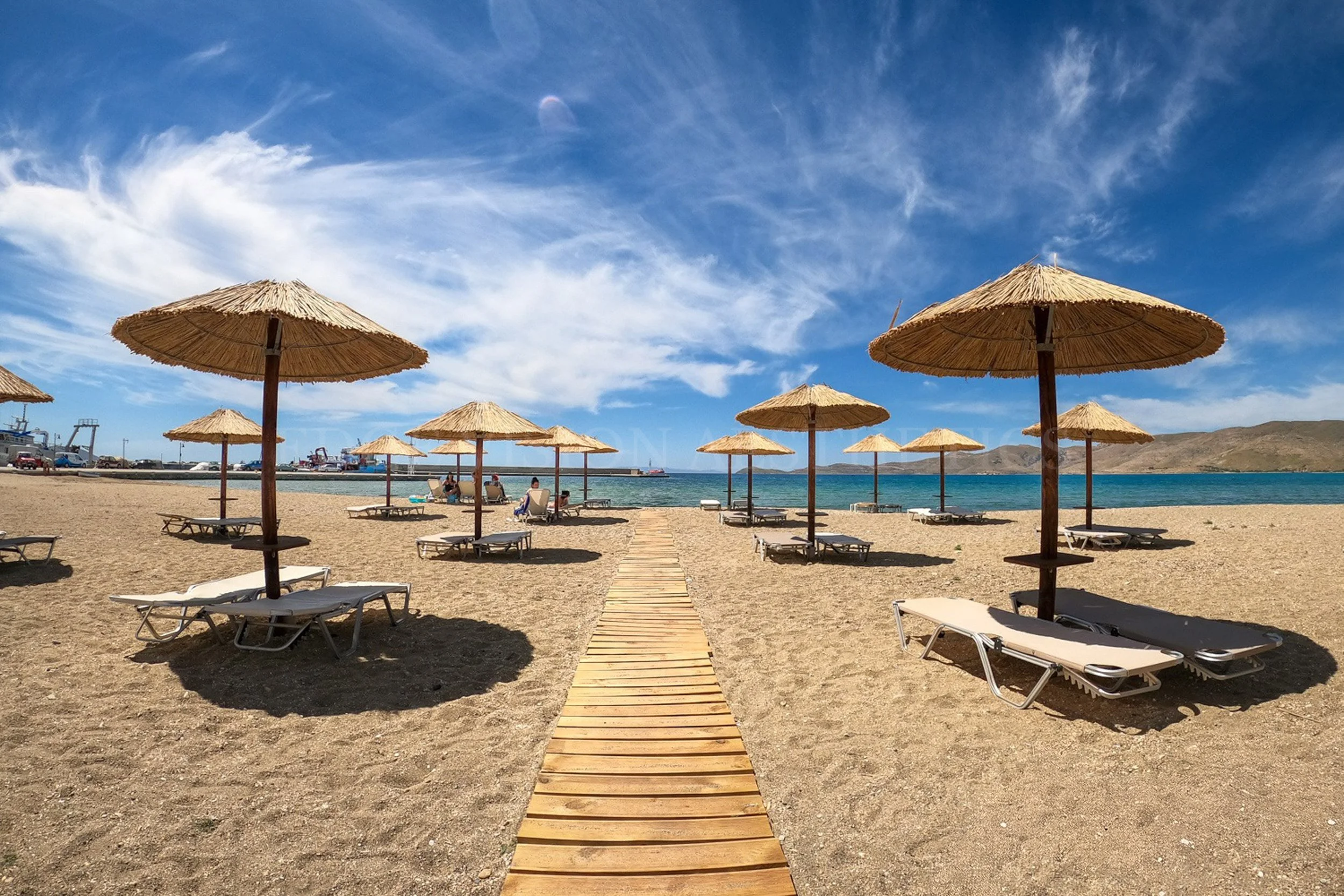 Beach with straw umbrellas, lounge chairs, and a wooden walkway leading to the water, with boats and hills in the background under a partly cloudy sky.