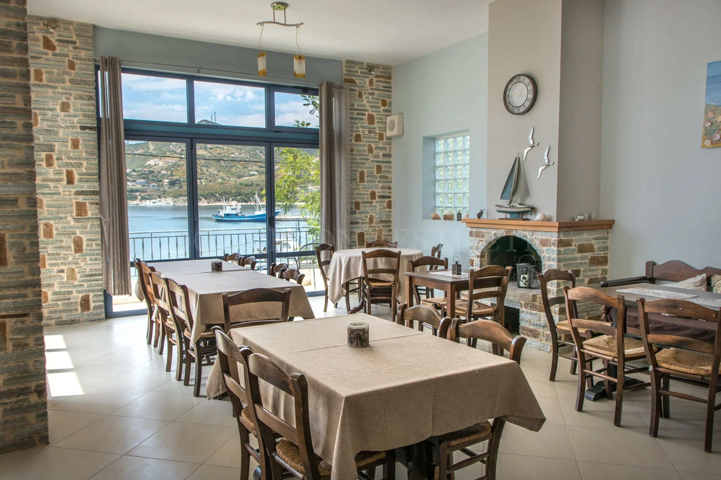 Interior of a restaurant with tables covered in beige tablecloths and wooden chairs, large windows showing a view of water and boats, stone and white walls, and decorative items including a clock and a model sailboat.