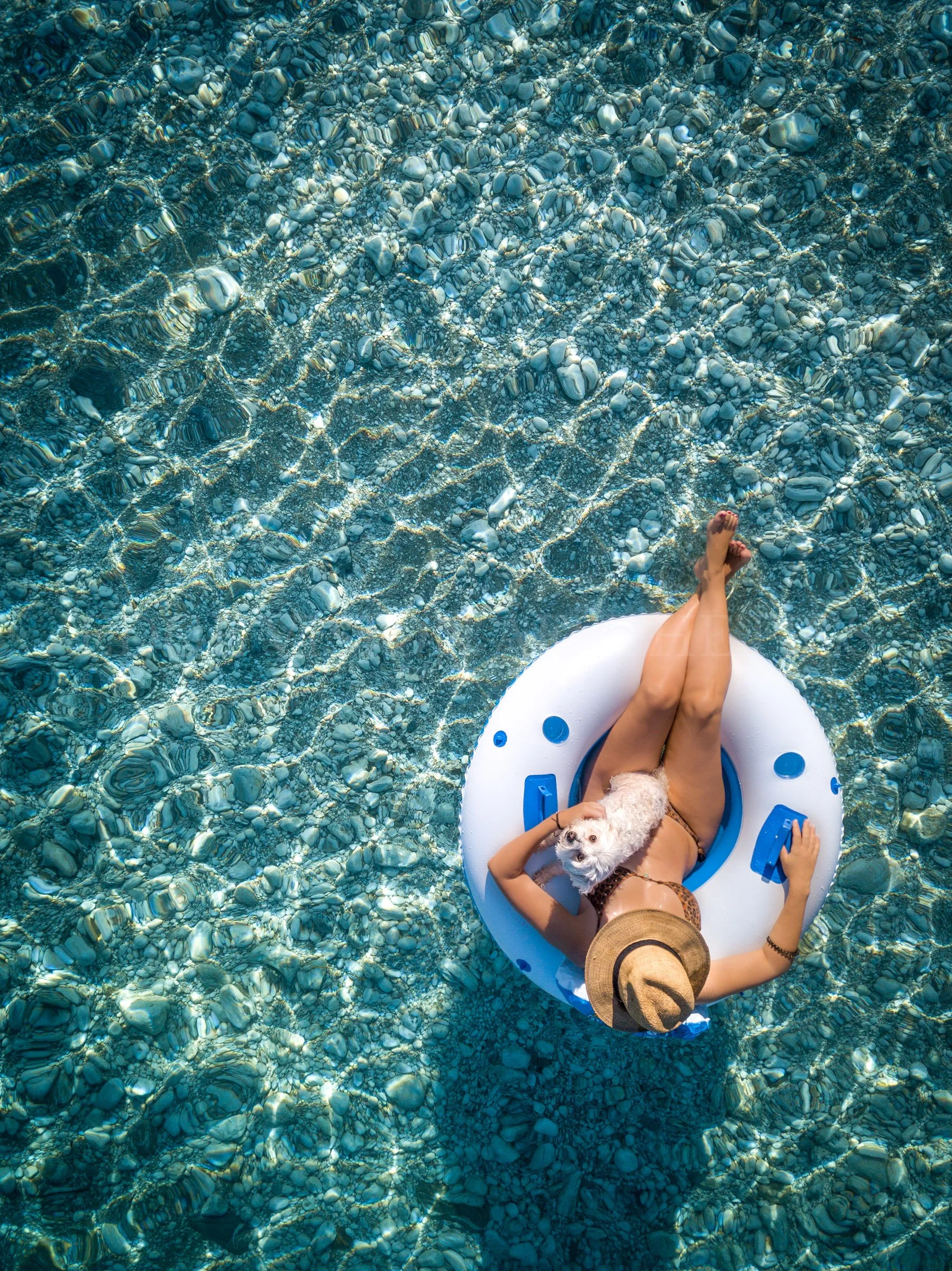 A person relaxing on a white inflatable float with blue accents in clear, rocky water, holding a small white dog, with the person's face partly visible and wearing a straw hat.