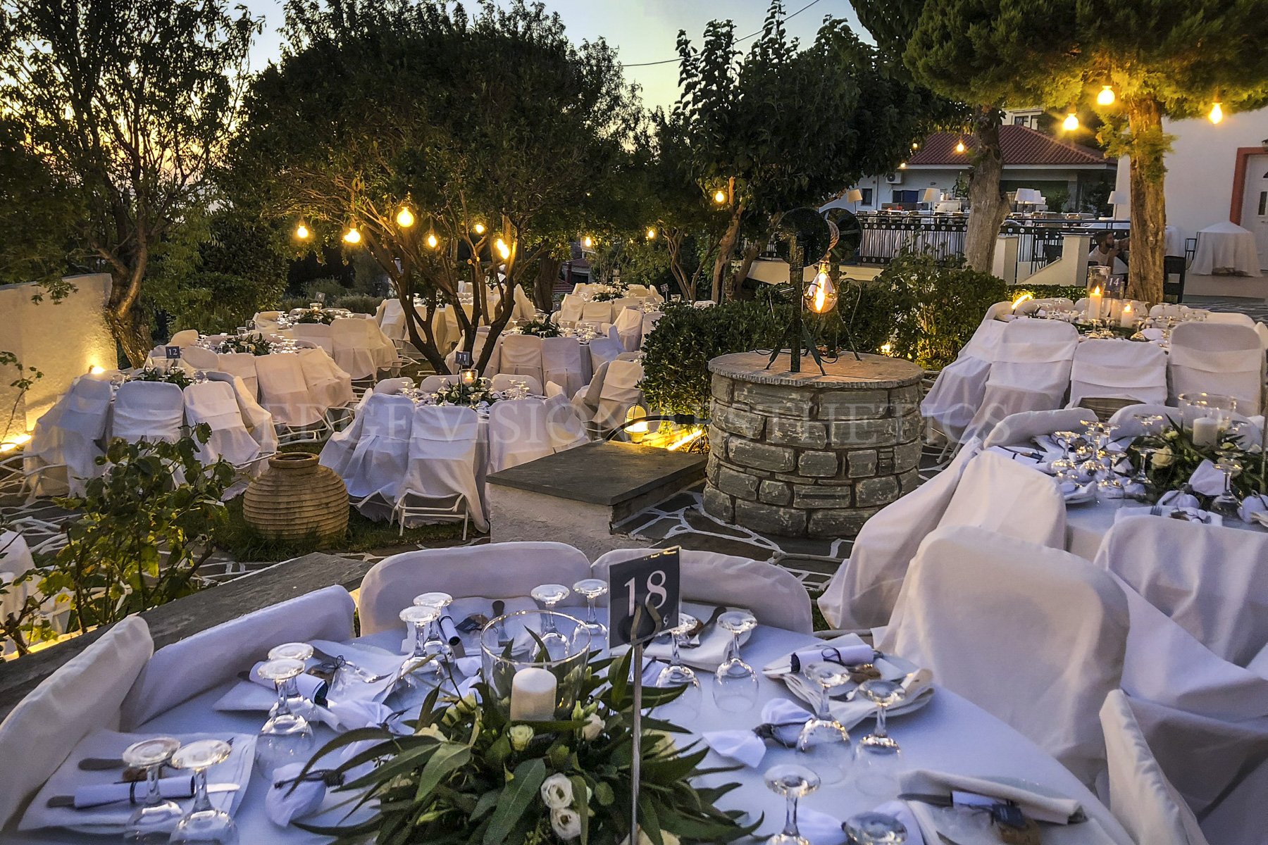 Outdoor dining area with round tables covered in white tablecloths, decorated with floral centerpieces and candles, set for a formal event during dusk. String lights hang overhead among trees, creating a warm ambiance, while chairs are covered in whi