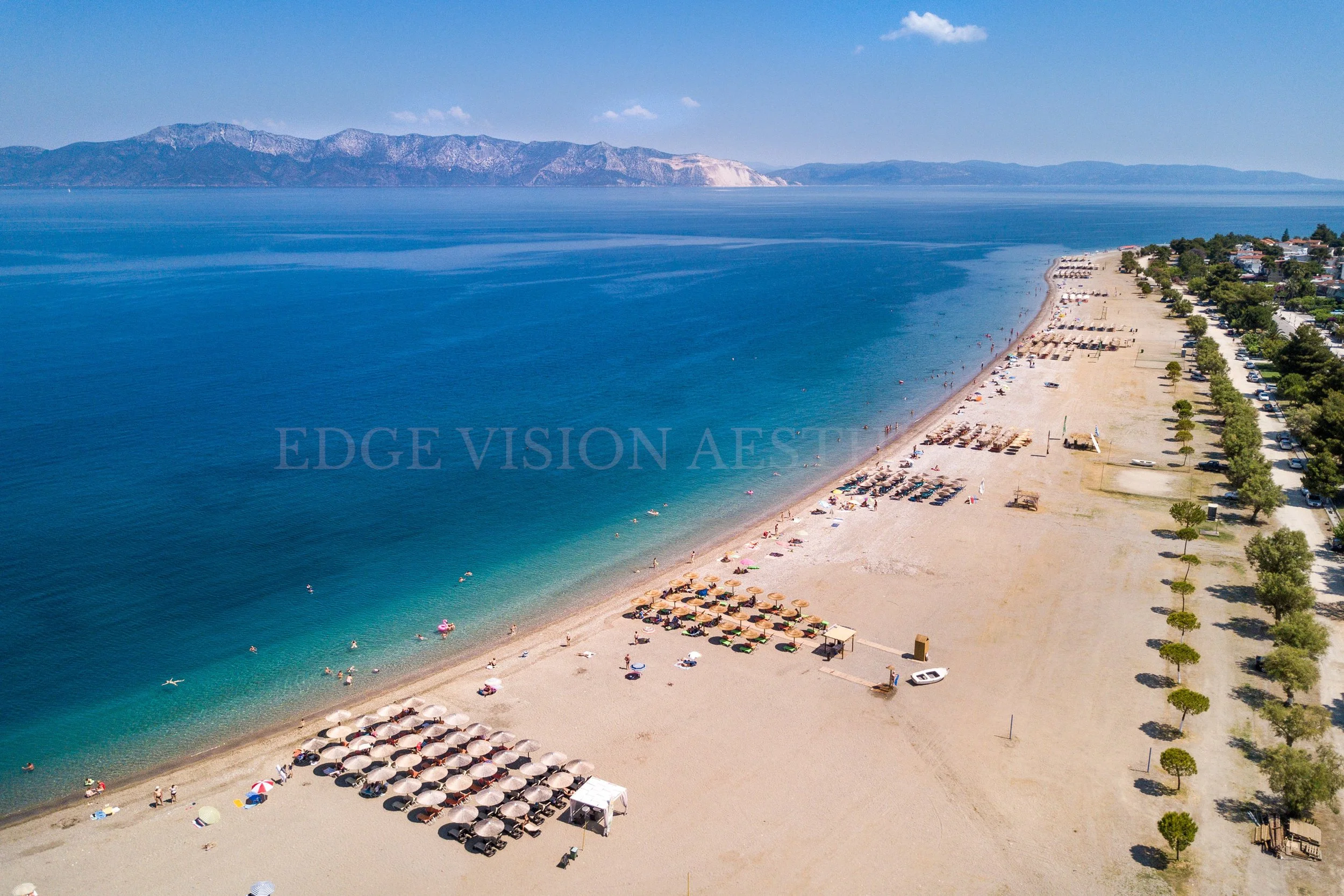 Aerial view of a beach with umbrellas and people, alongside a strip of trees and parked cars, with mountains in the background and a blue sky with few clouds.
