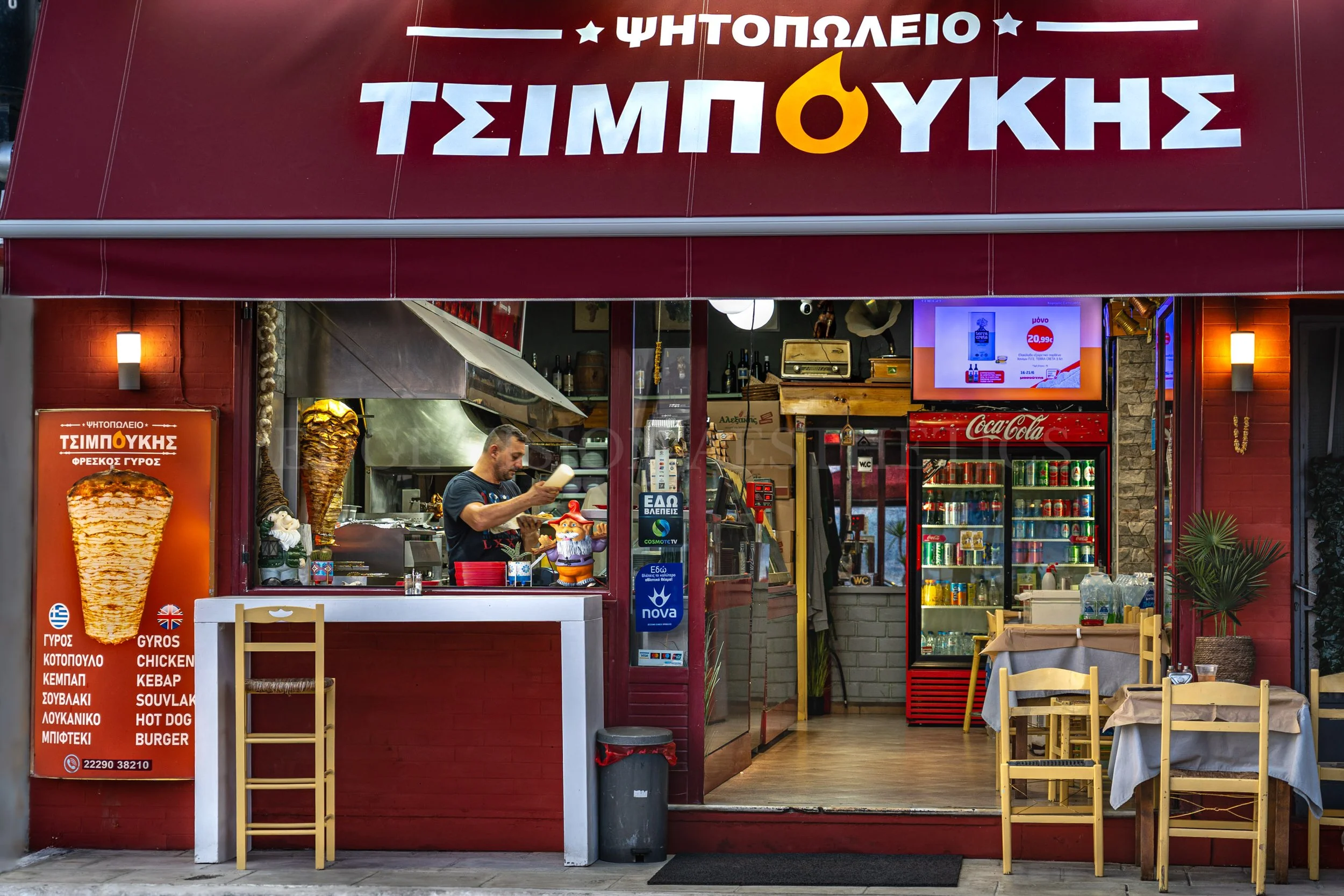Front view of a Greek gyros shop with red signage, menu in Greek and English, and a man preparing food inside. There are outdoor chairs and tables for customers.