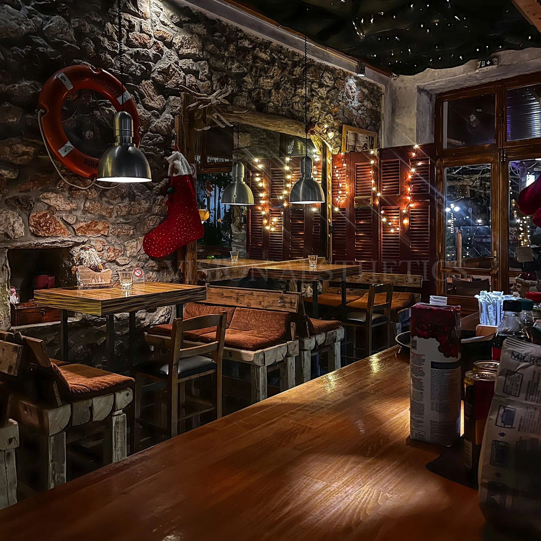 A cozy restaurant interior decorated with a rustic stone wall, fairy lights, and a Christmas stocking hanging near a fireplace. There are wooden tables and chairs, with some tables set with glasses. The ambiance is warm and festive, with Christmas de