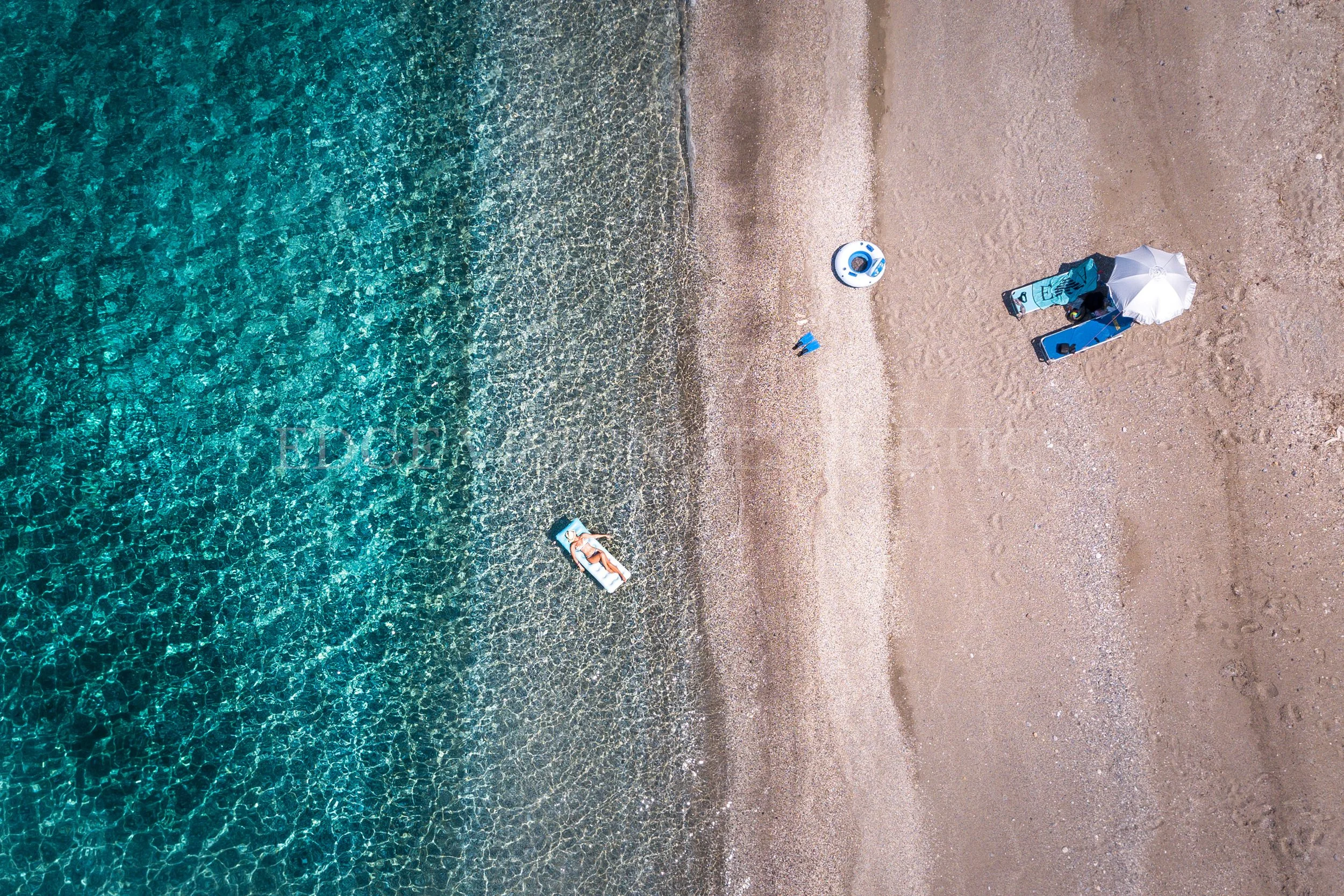 Aerial view of a beach showing clear blue water, sandy shore, and several people relaxing under umbrellas.