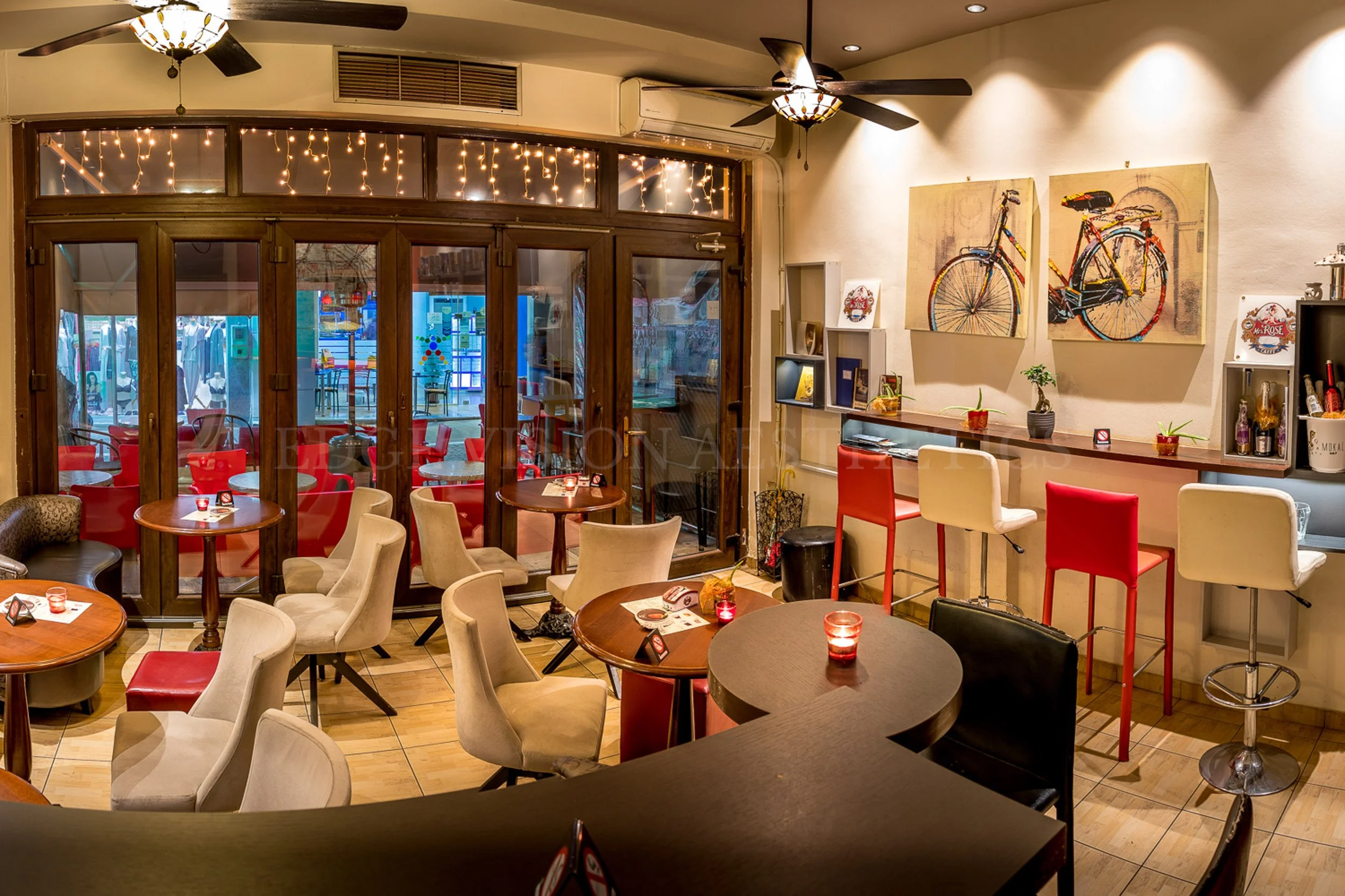 A cozy indoor restaurant or cafe with round wooden tables, some with red candles, surrounded by beige upholstered chairs and a few red and white barstools. The back wall has two bike paintings, shelves with books, plants, and bottles, and is decorate