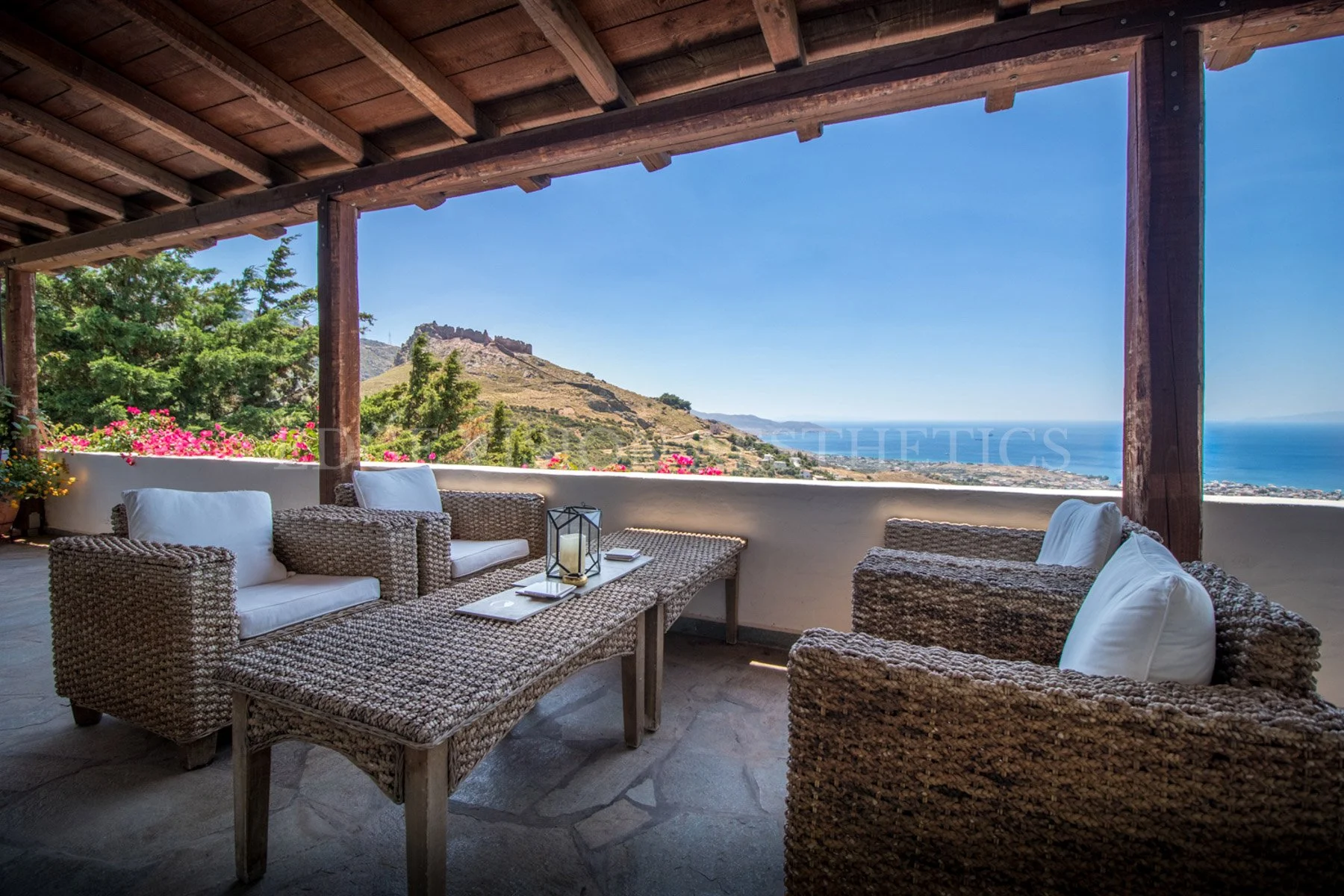Outdoor patio with woven furniture, pillows, and lanterns, overlooking hills and ocean under a clear blue sky.