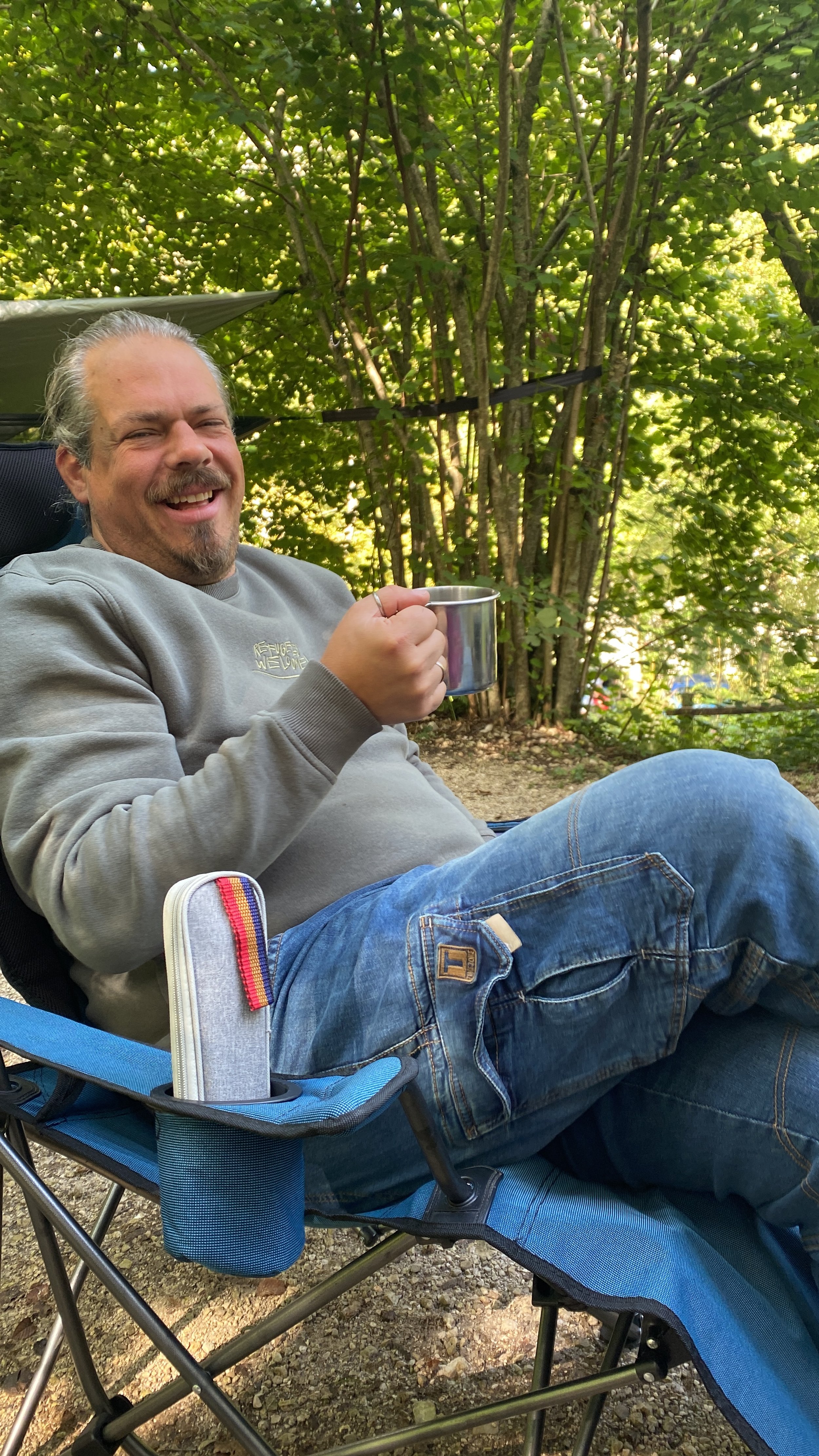 Mann sitzt lächelnd auf einem Campingstuhl im Freien mit einem Metallbecher in der Hand, umgeben von Bäumen.