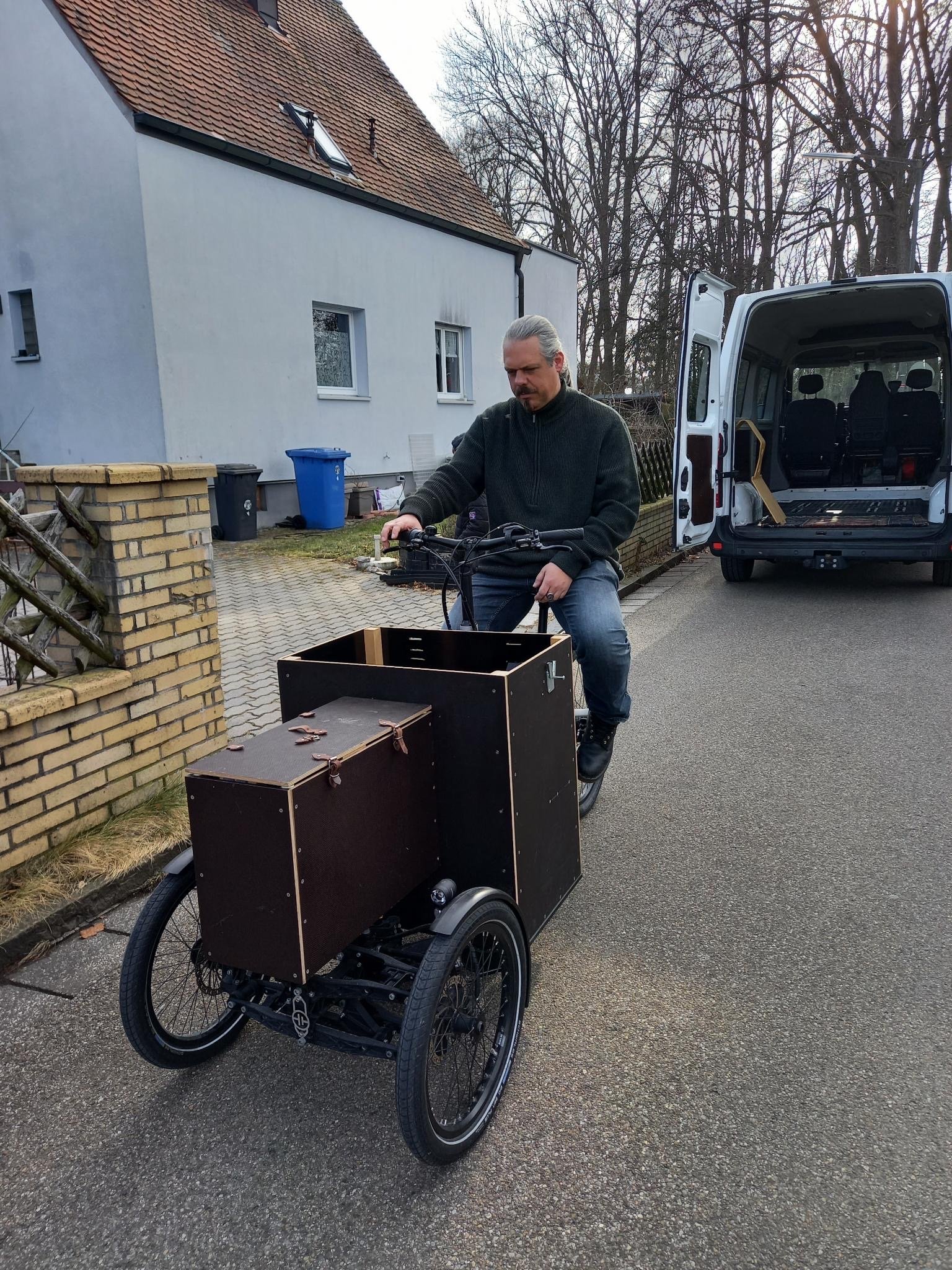 Ein Mann fährt auf einem dreirädrigen Lastenfahrrad auf einer Straße vor einem Haus mit roten Dachziegeln. Im Hintergrund steht ein geparkter, geöffneter Lieferwagen. Bäume ohne Blätter sind im Hintergrund sichtbar.