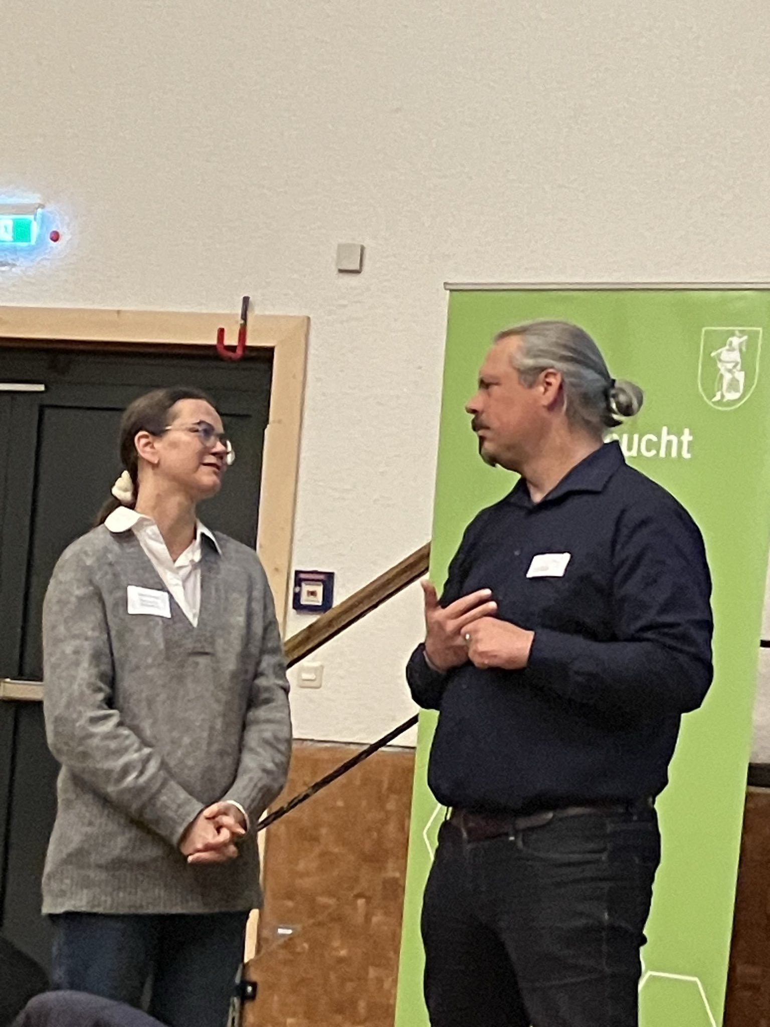 Zwei Personen in einem Gespräch; die Frau links trägt einen grauen Pullover, der Mann rechts ein dunkles Hemd. Im Hintergrund ist ein grünes Banner. Sie befinden sich in einem Innenraum.