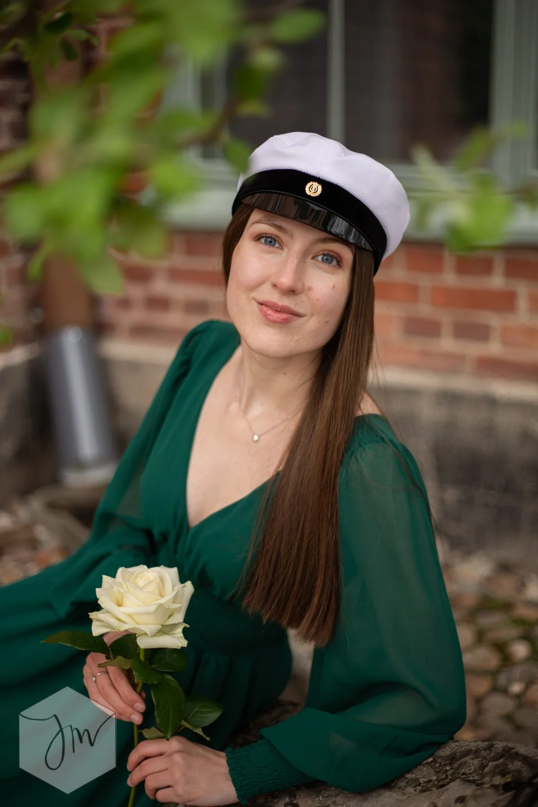 Nuori nainen pukeutuneena vihreään mekkoon, päässään ylioppilaslakki. Istuu ulkona pienen puunoksan päällä, pitelee valkoista ruusua kädessään ja hymyilee kameralle.