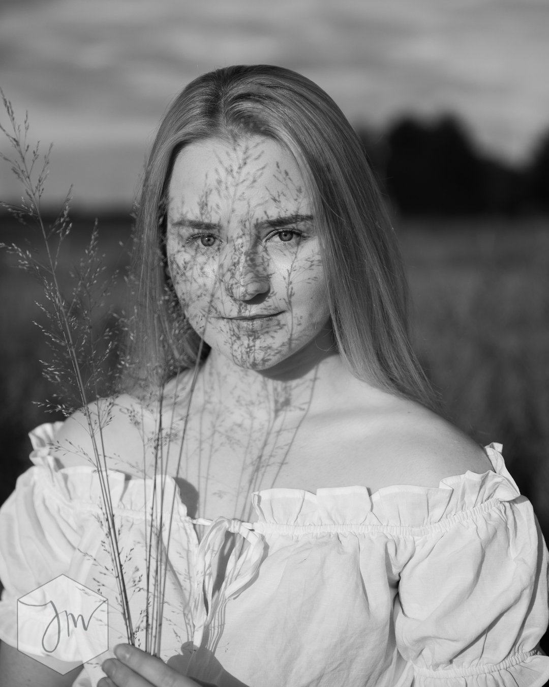 Mustavalkovalokuva nuoresta naisesta, joka seisoo ulkona ja katsoo kameraan. Hänen kasvonsa on osittain varjostettu heinällä, joka on hänen edessään. Taustalla on peltoa ja pilvinen taivas.