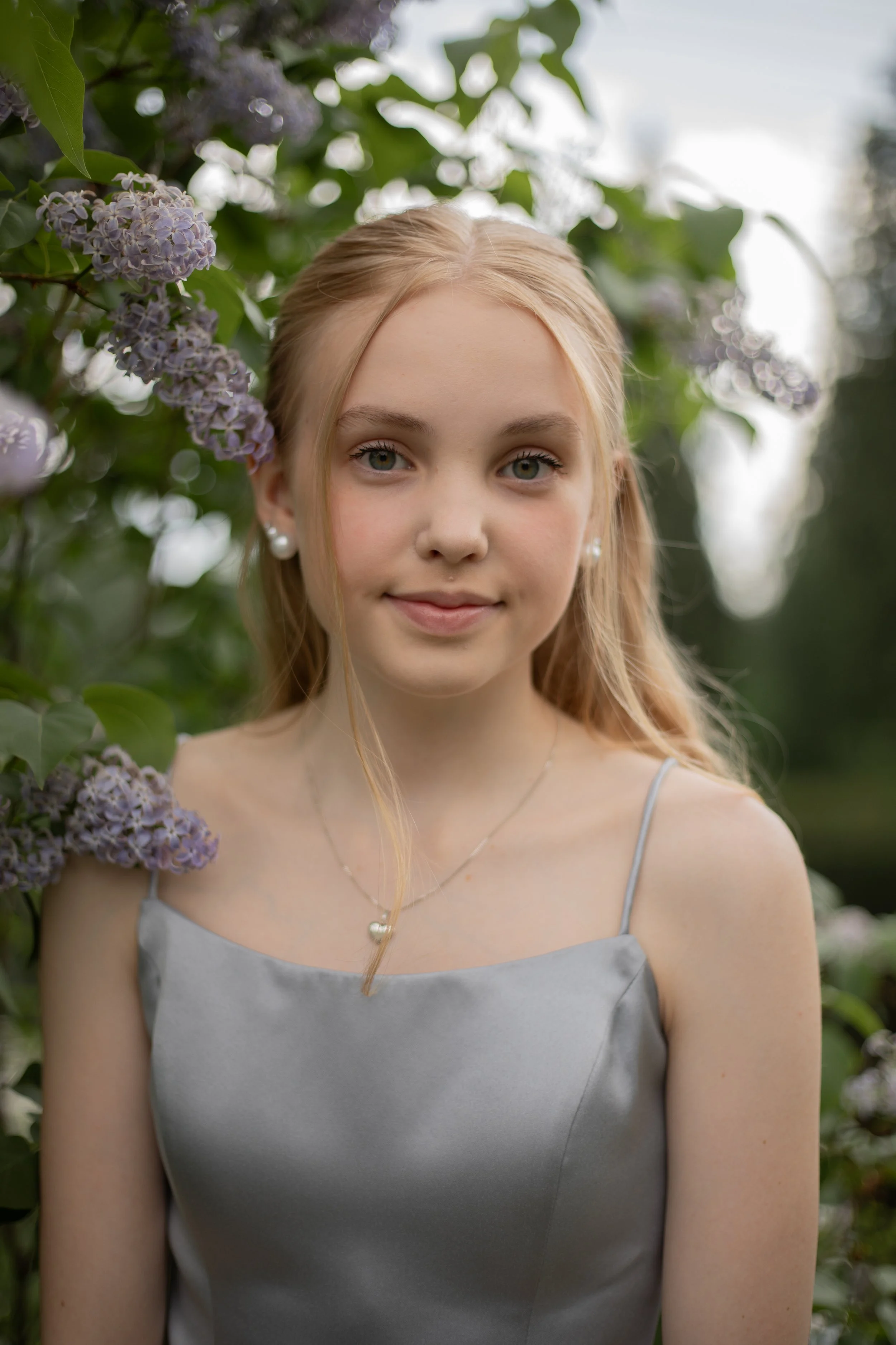 Nuori tyttö poseeraa puistossa, taustalla vihreitä lehtiä ja kukkia, hänellä on hopeanvärinen mekko ja korvakorut.