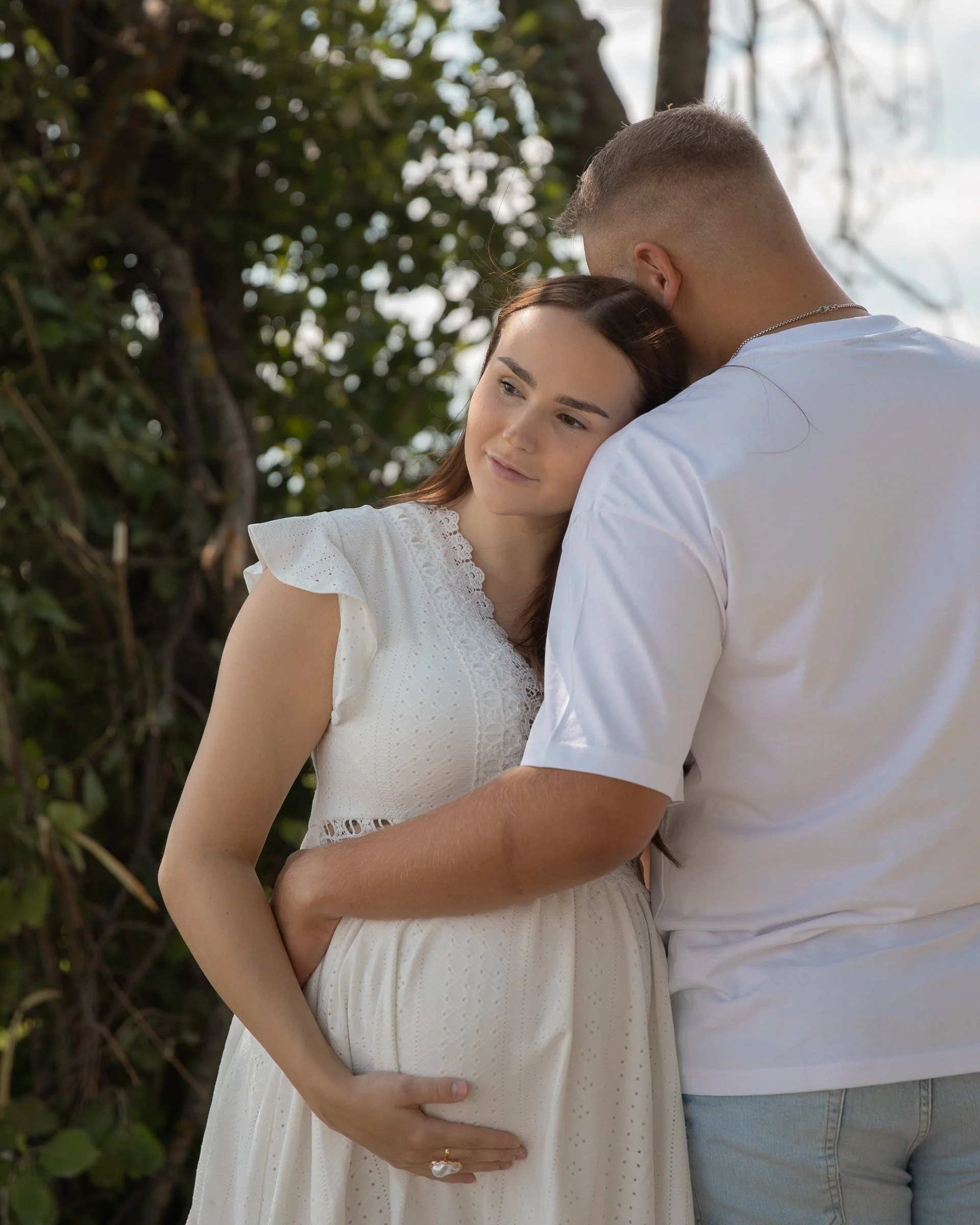 Raskaana oleva nainen ja mies halaavat ulkona, taustalla puut ja taivasta