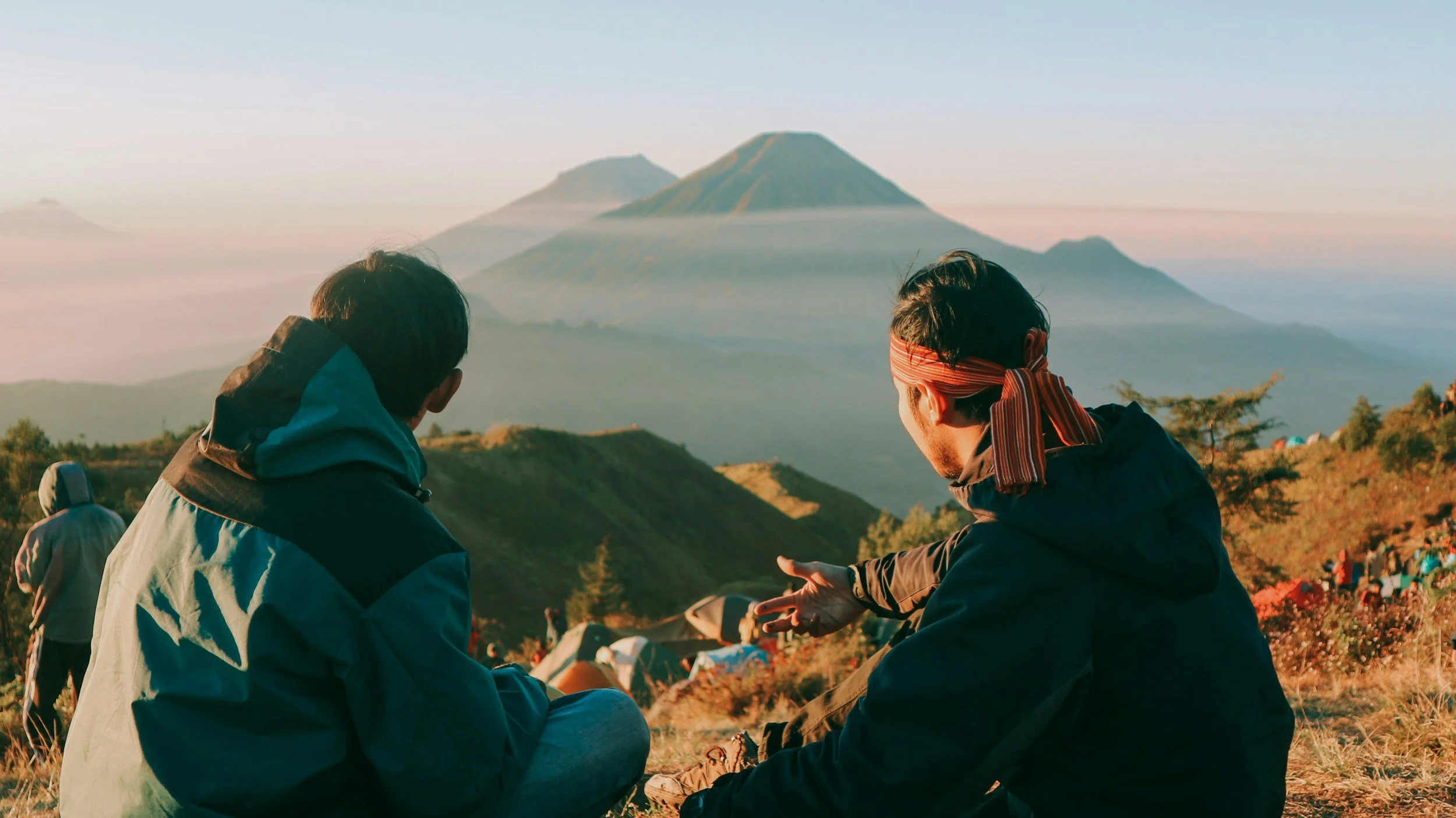 Two people sitting on a hill overlooking a mountain range, with tents and vegetation in the foreground.