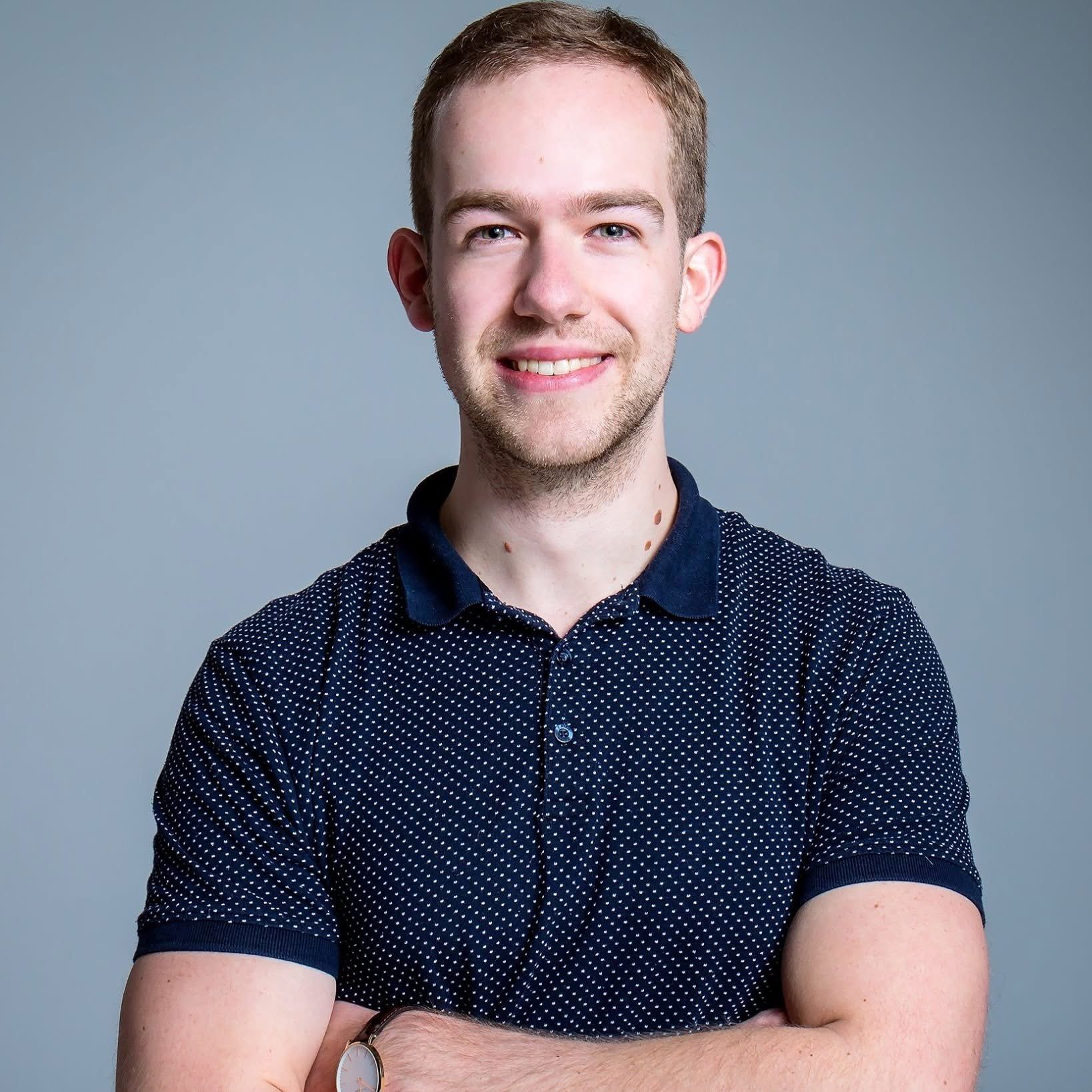 Man in a dark blue polka dot shirt with folded arms, smiling, against a gray background.