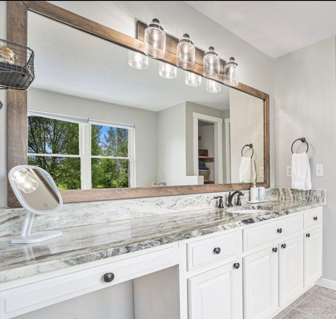 Modern bathroom vanity with large mirror, marble countertop, dual sinks, and pendant lights. The vanity area includes white cabinetry, towel rings with white towels, and a small magnifying mirror. Natural light enters from a window with a view of tre