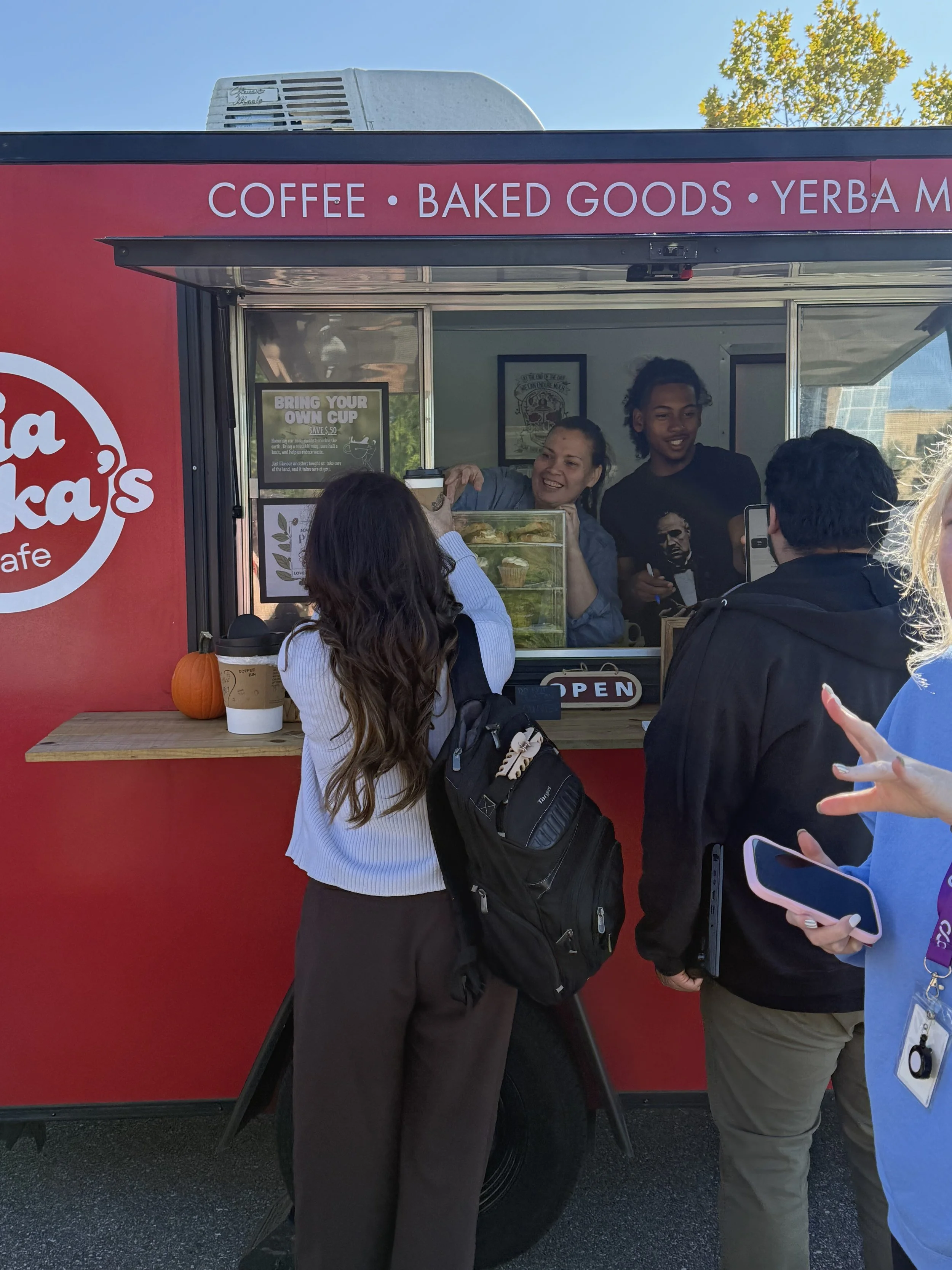 A group of people waiting in line at a food truck that offers coffee, baked goods, and Yerba Mate, with the truck's window open and two employees visible, one holding baked goods and smiling.