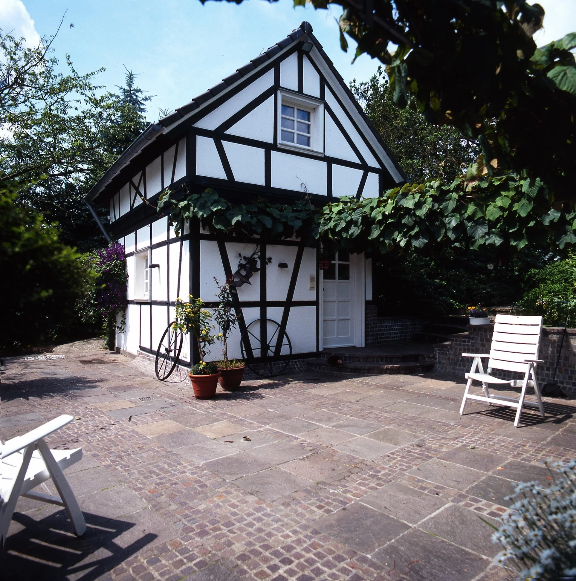 Die Remise des komplett sanierten Fachwerkhauses, umgeben von Gartenpflanzen, auf gepflastertem Hof mit Gartenmöbeln.